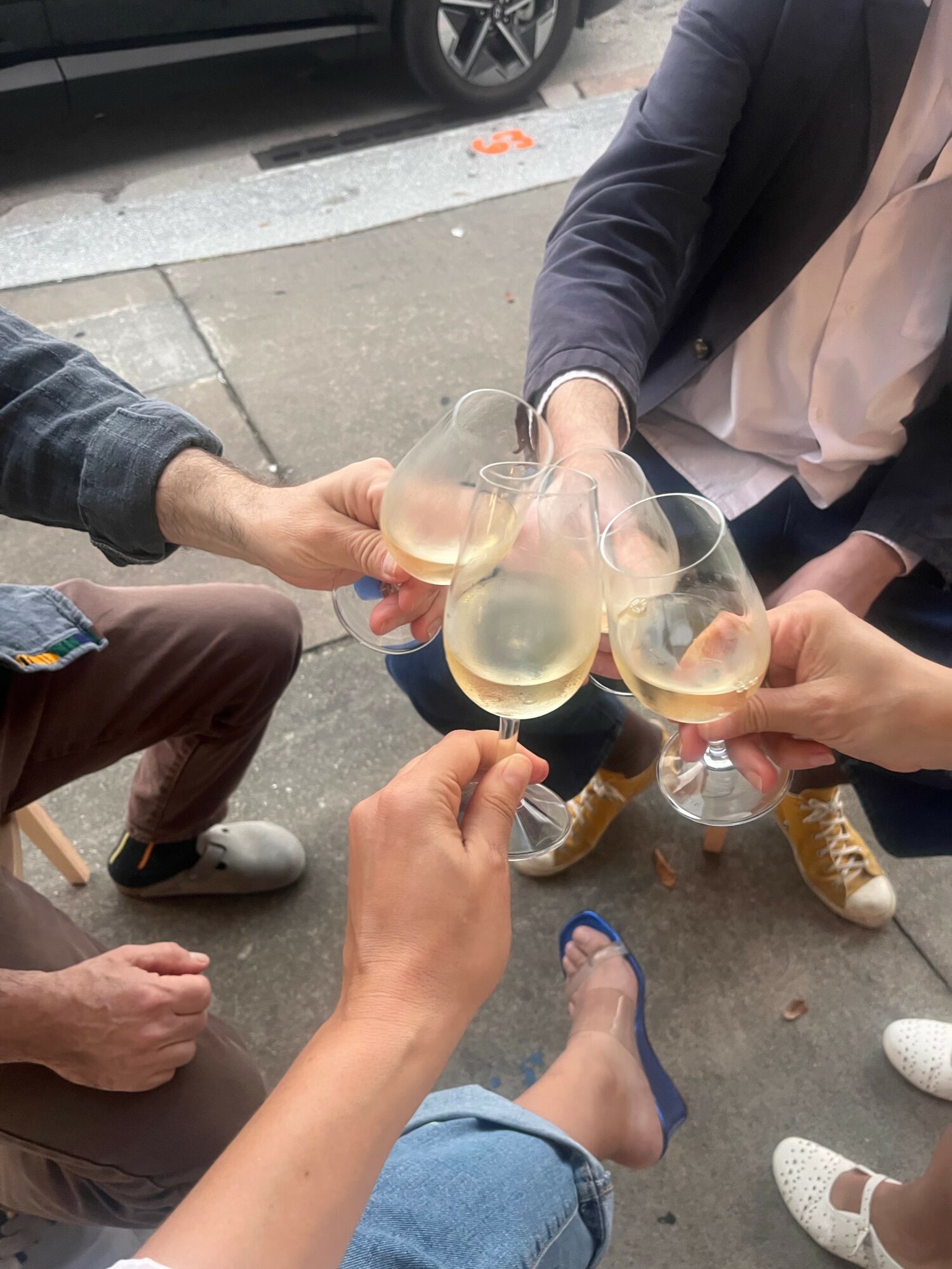 Four people clink glasses of champagne outdoors on a sidewalk, with a car and street visible in the background.