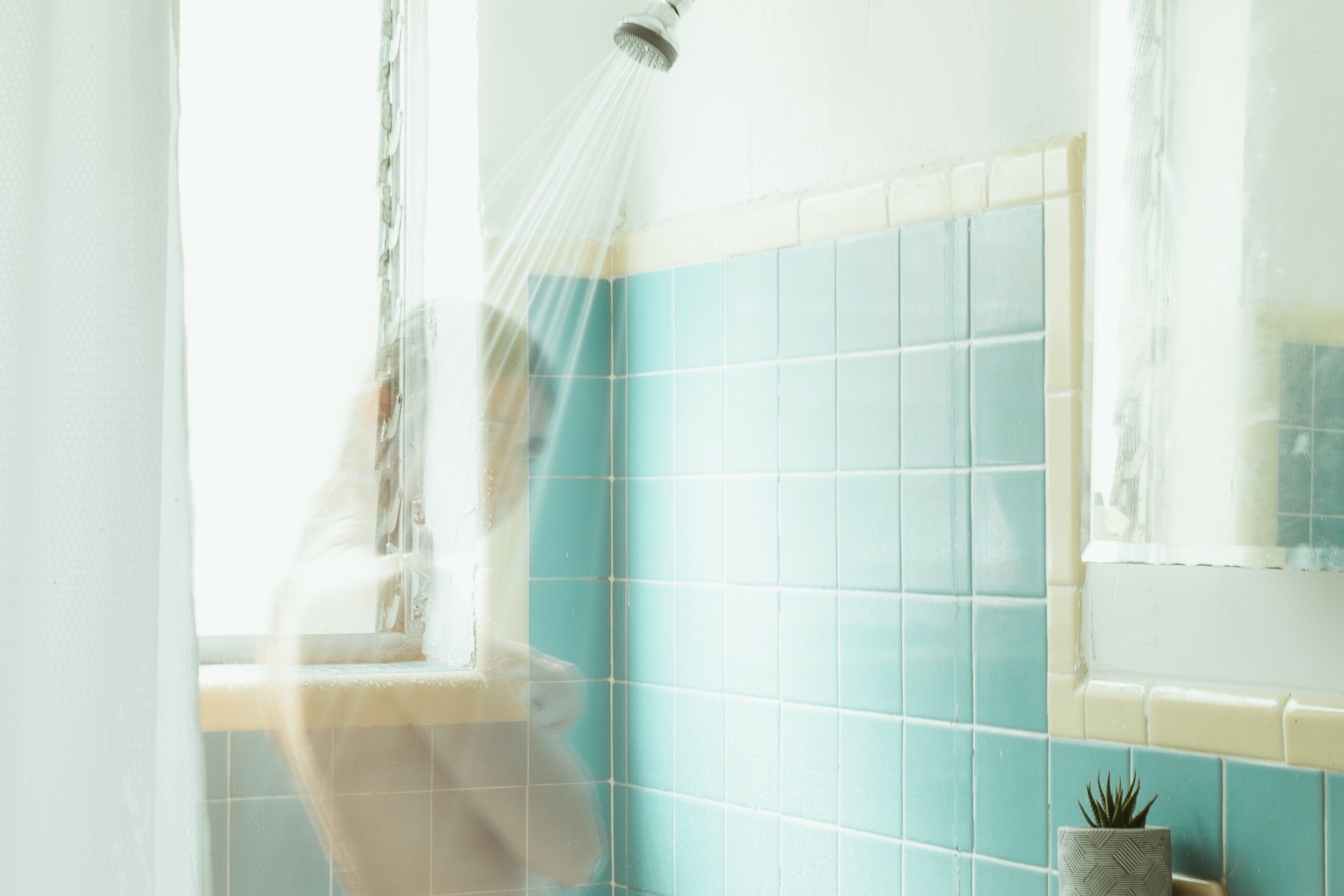 Shower with water spray, window, and tiled wall, with a person partially visible behind the curtain.