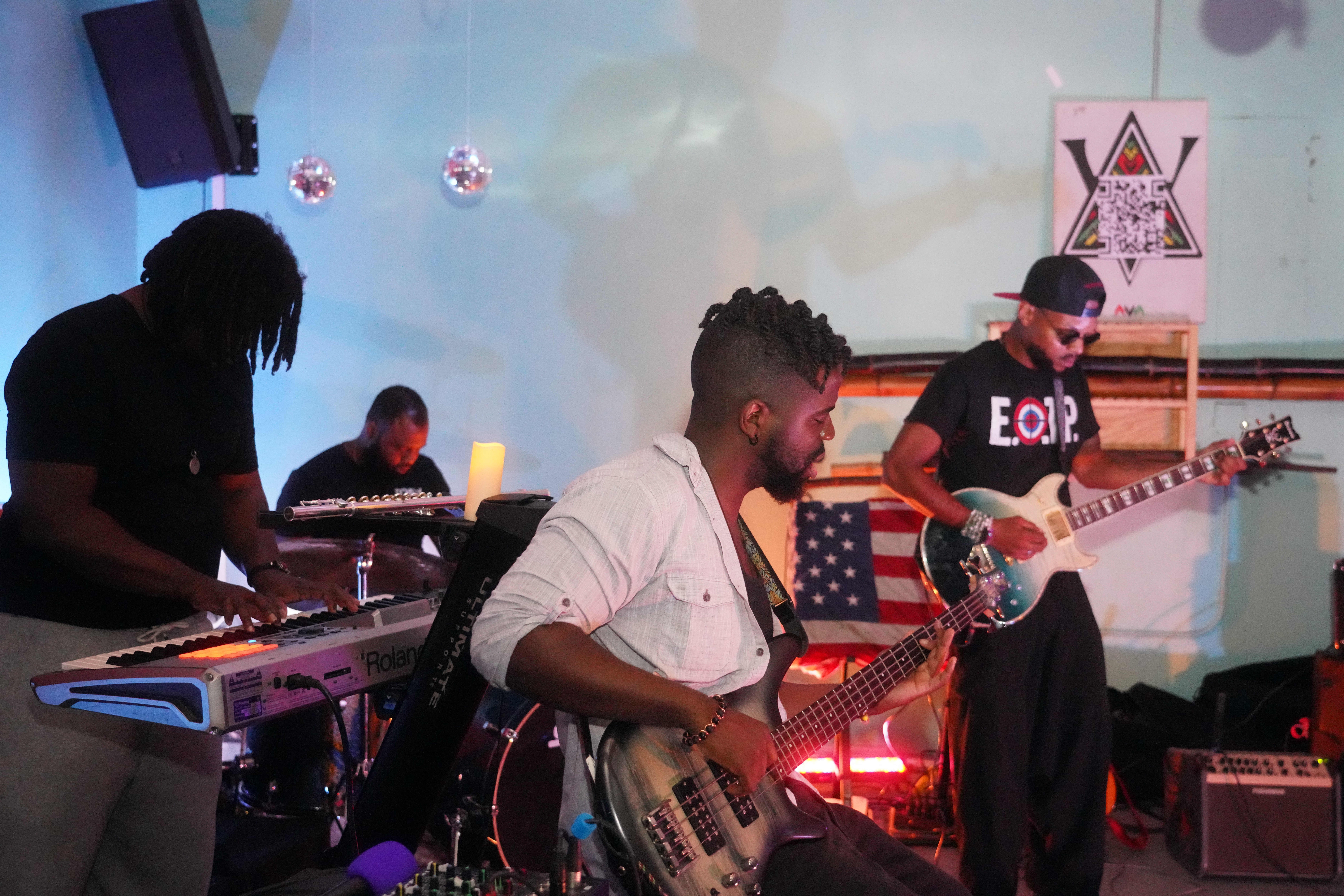 Four musicians perform on stage with instruments, including a keyboard, bass guitar, and electric guitar, in a dimly lit setting.
