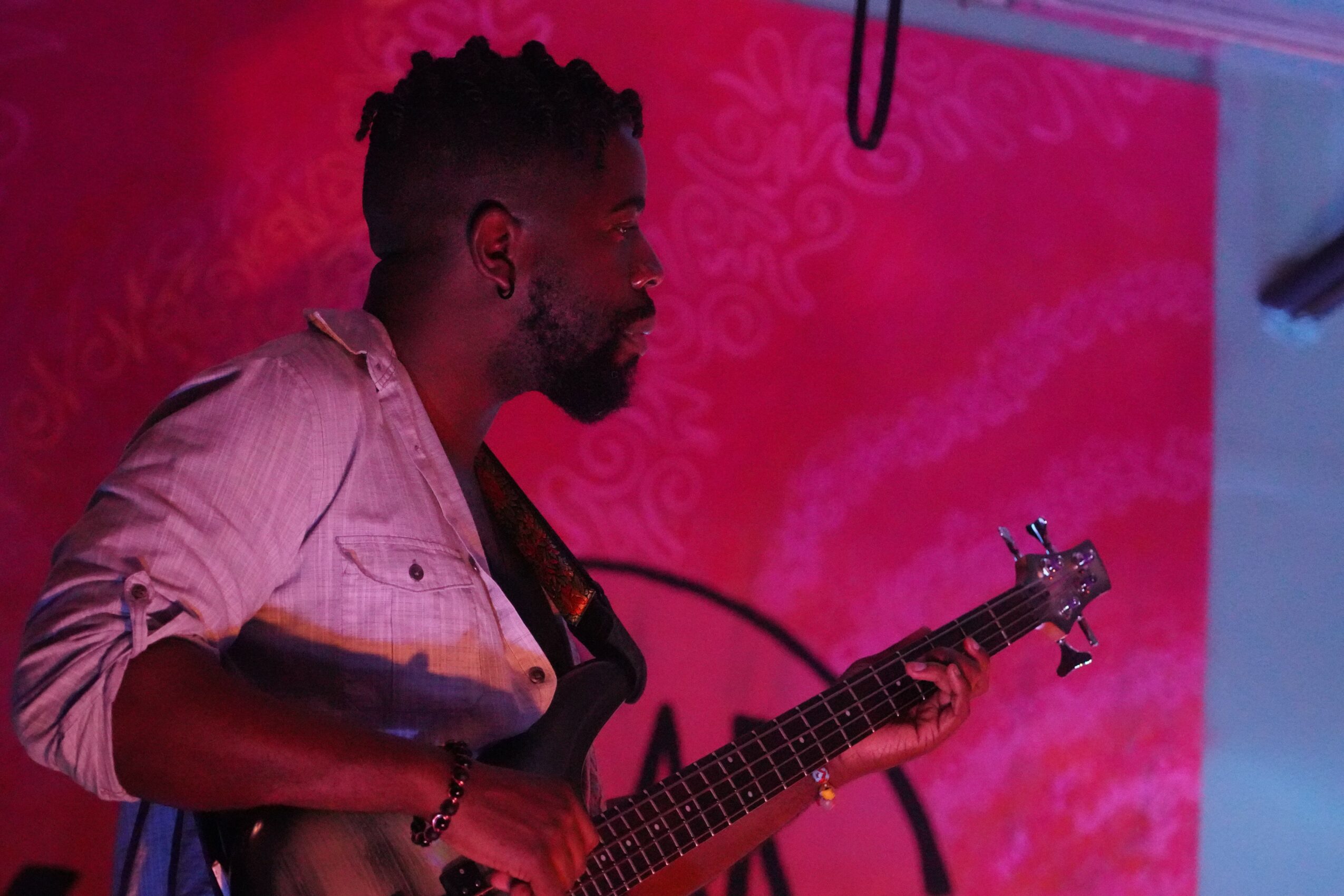 Man playing guitar on stage with pink background, side profile, wearing light-colored shirt.