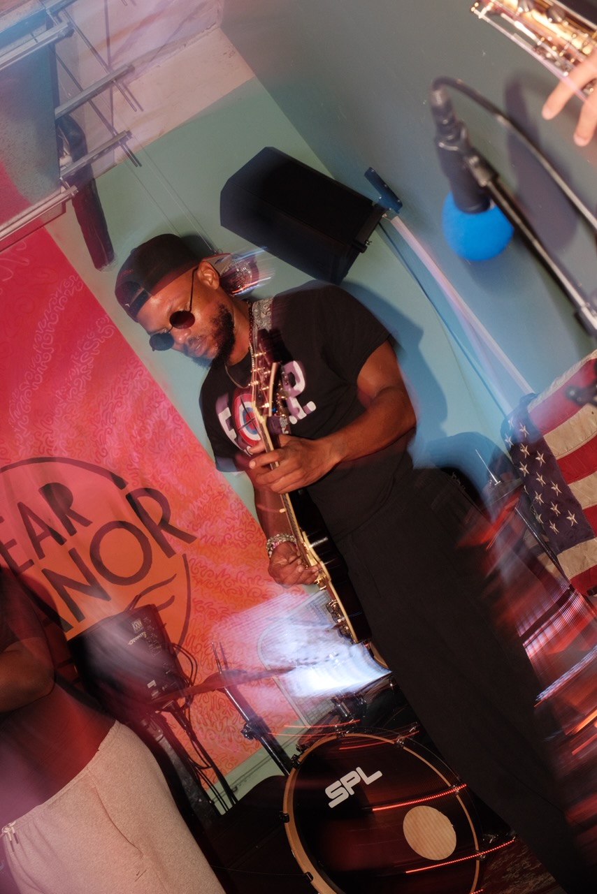 Person wearing sunglasses and a cap, holding a microphone, performing on stage with a colorful background.