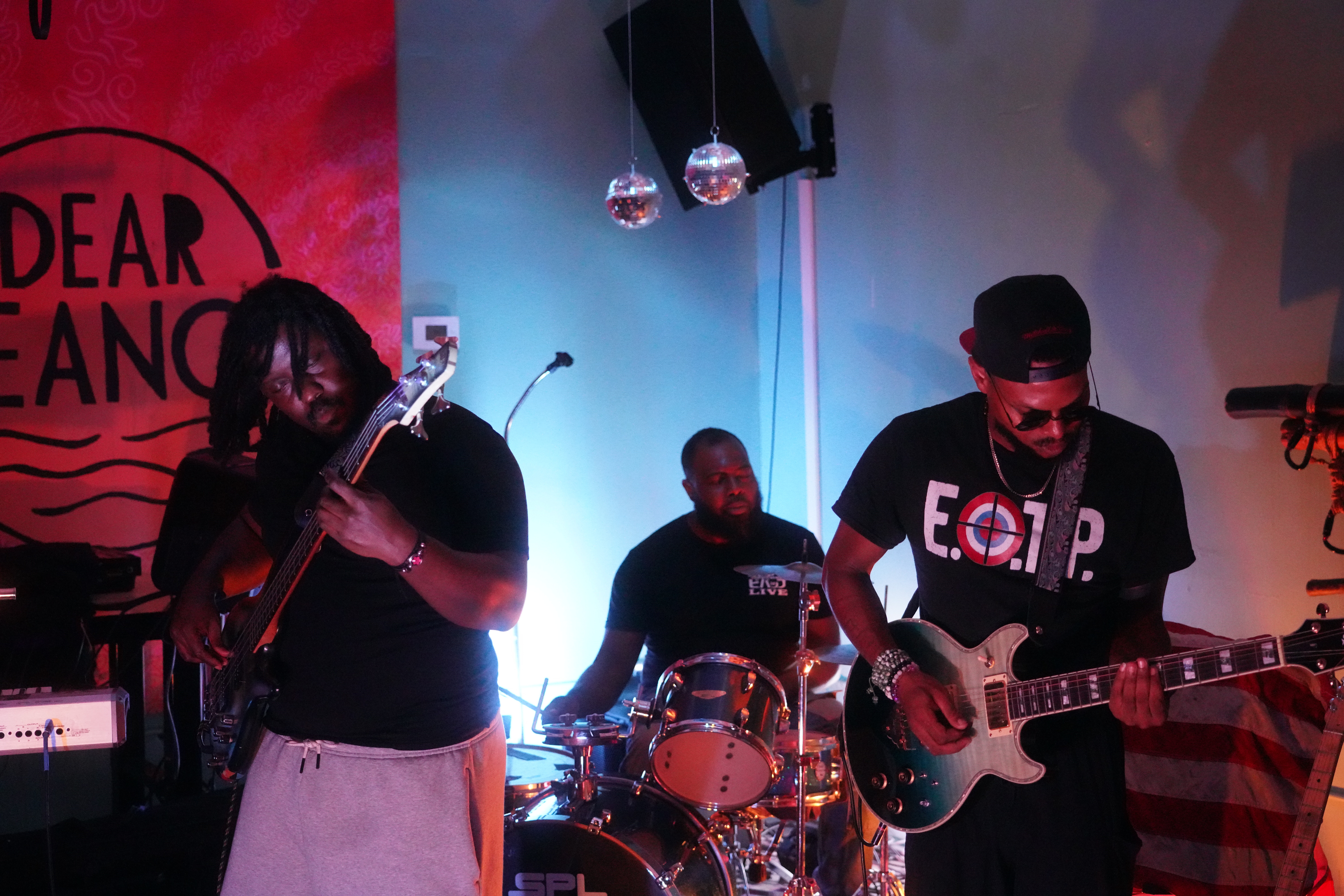 Three musicians perform on stage with instruments, a drum set, and a backdrop with text, under colorful lighting.