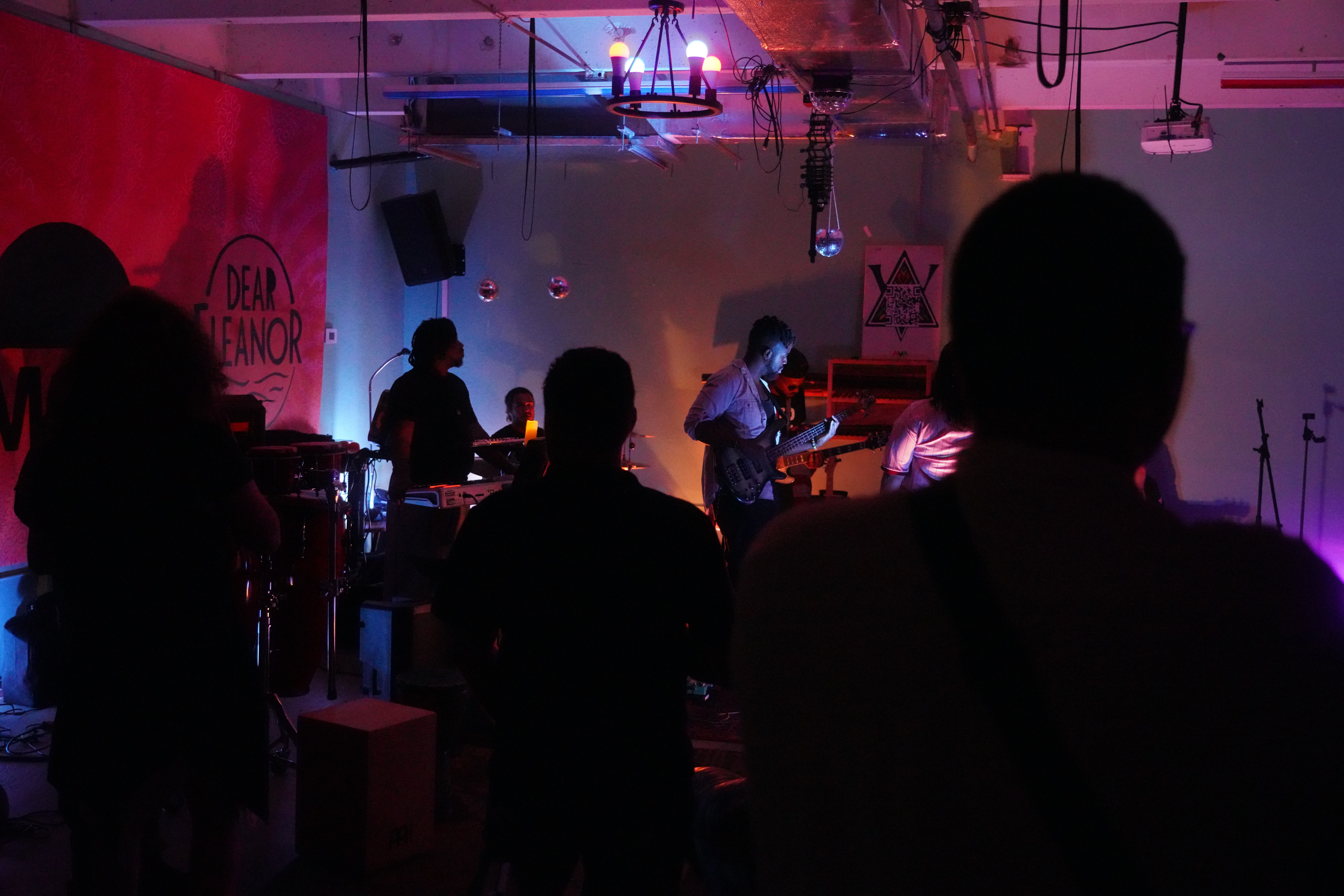 Band performing on stage with audience watching in a dimly lit venue, colorful lighting, and musical equipment visible.