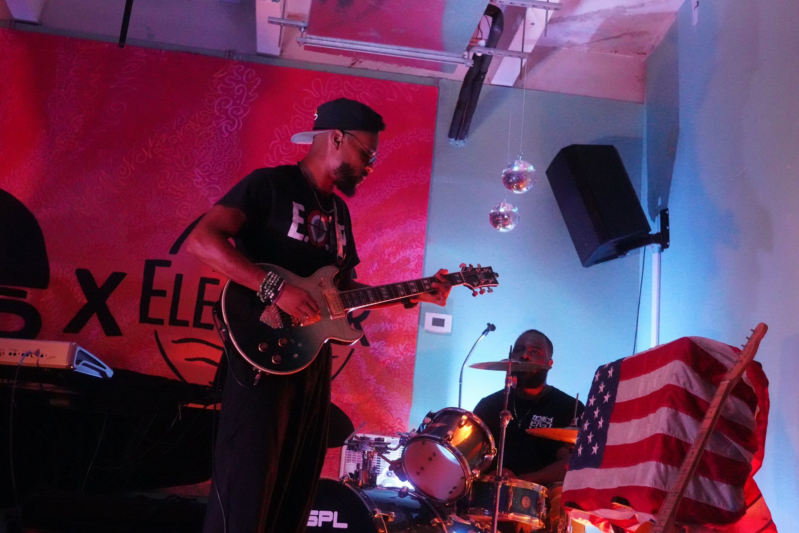 Musician playing guitar on stage with a drummer, American flag, and stage lighting, in a performance setting.