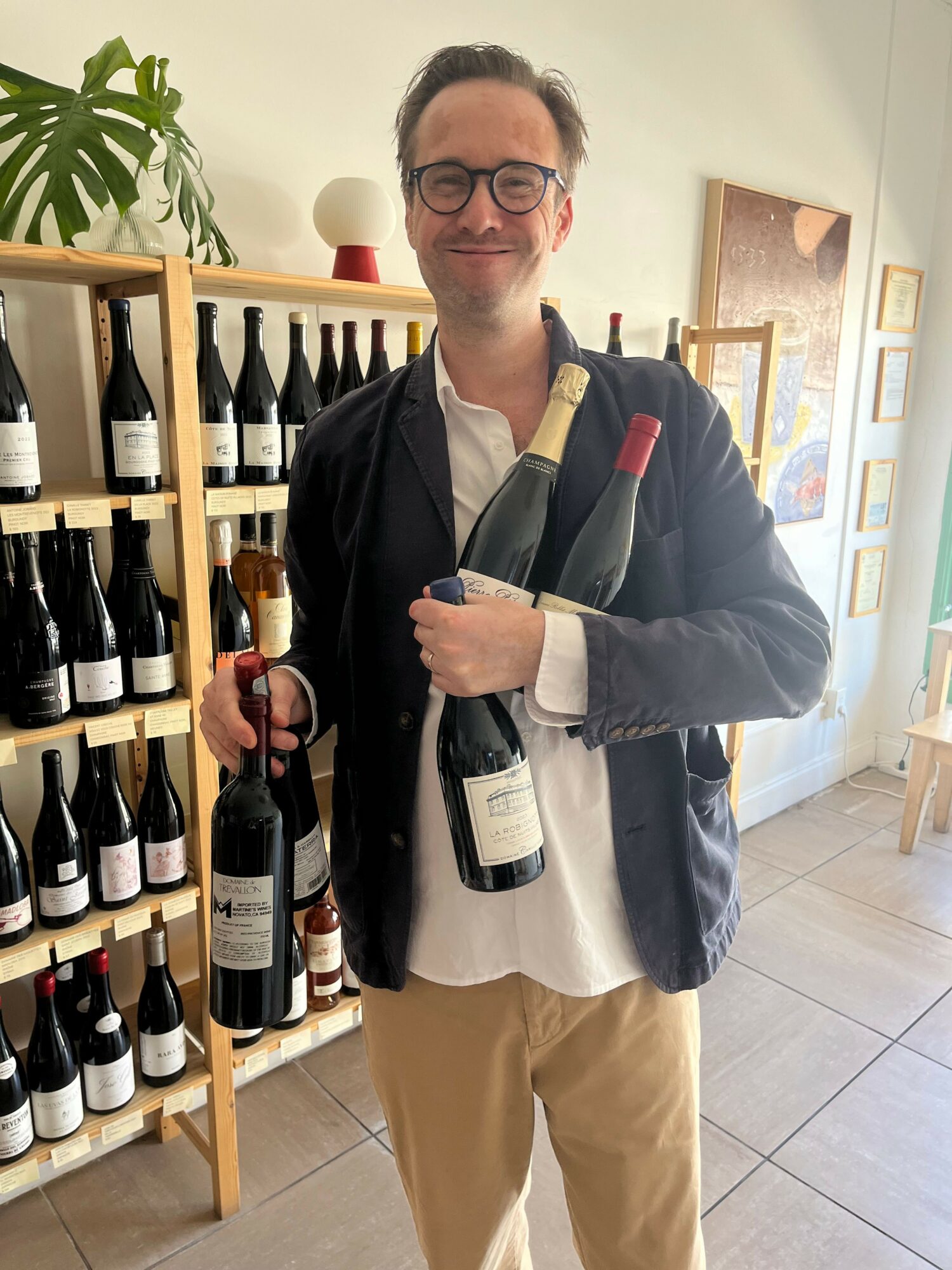 Man holding a wine bottle, standing indoors with a wine rack and artwork in the background.