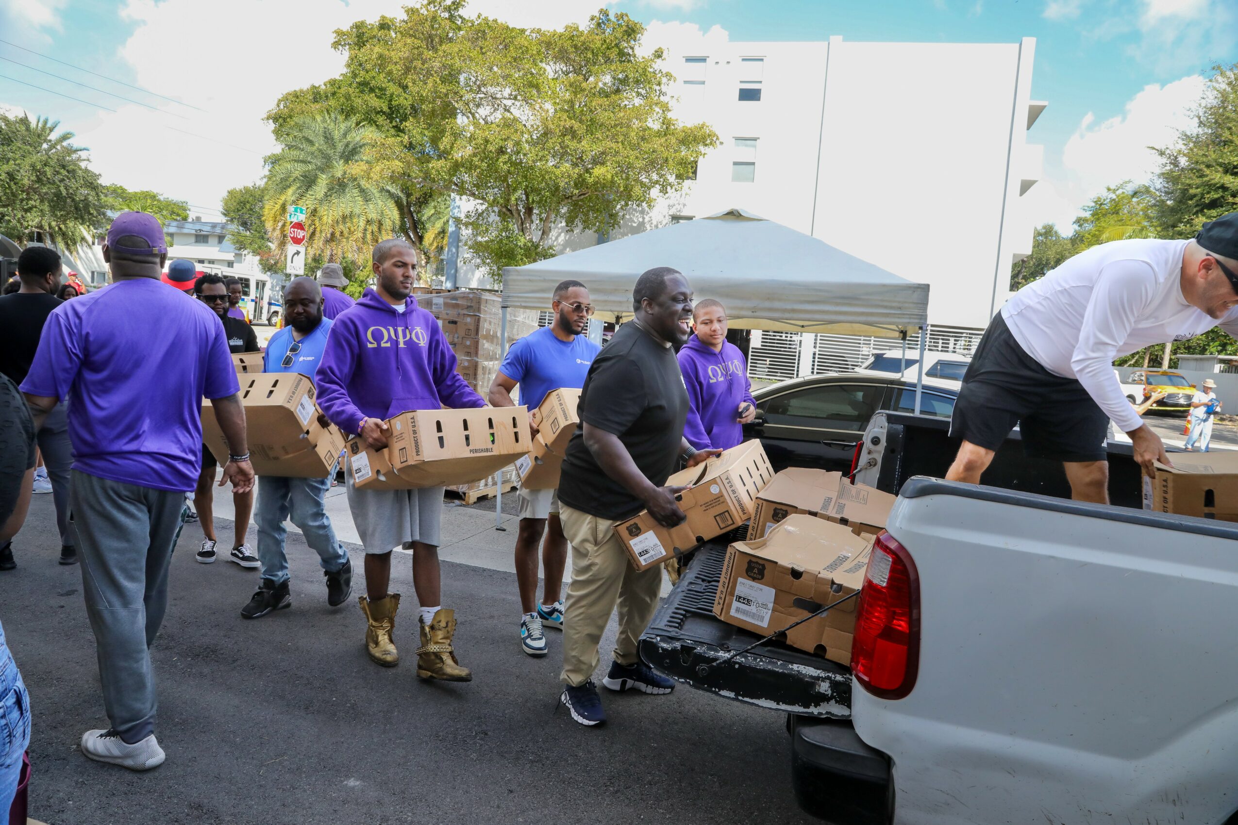 Group of people unloading boxes from a pickup truck outdoors, with trees and buildings in the background.