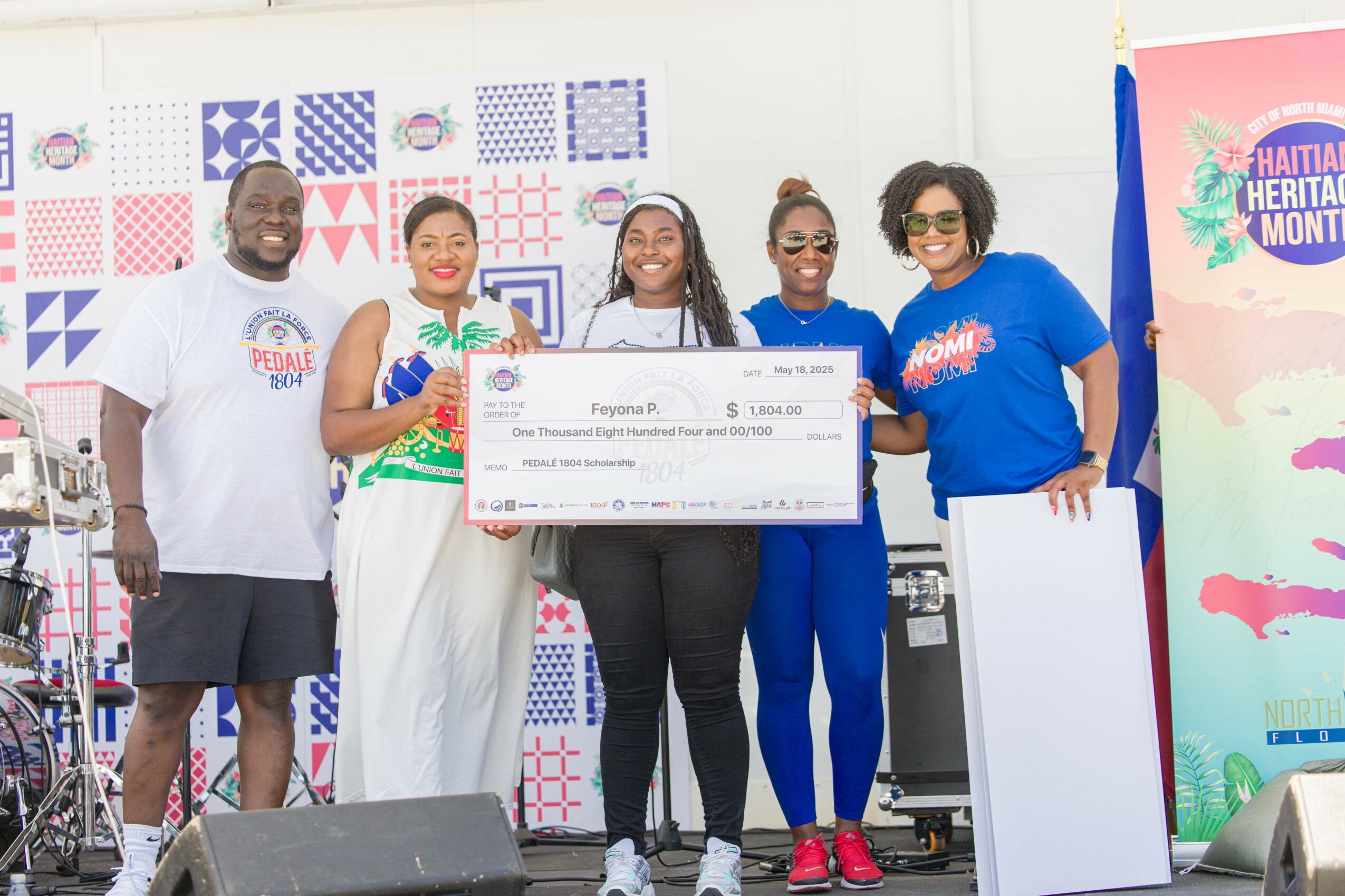 Group of five people holding a large check at an outdoor event, with colorful banners in the background.