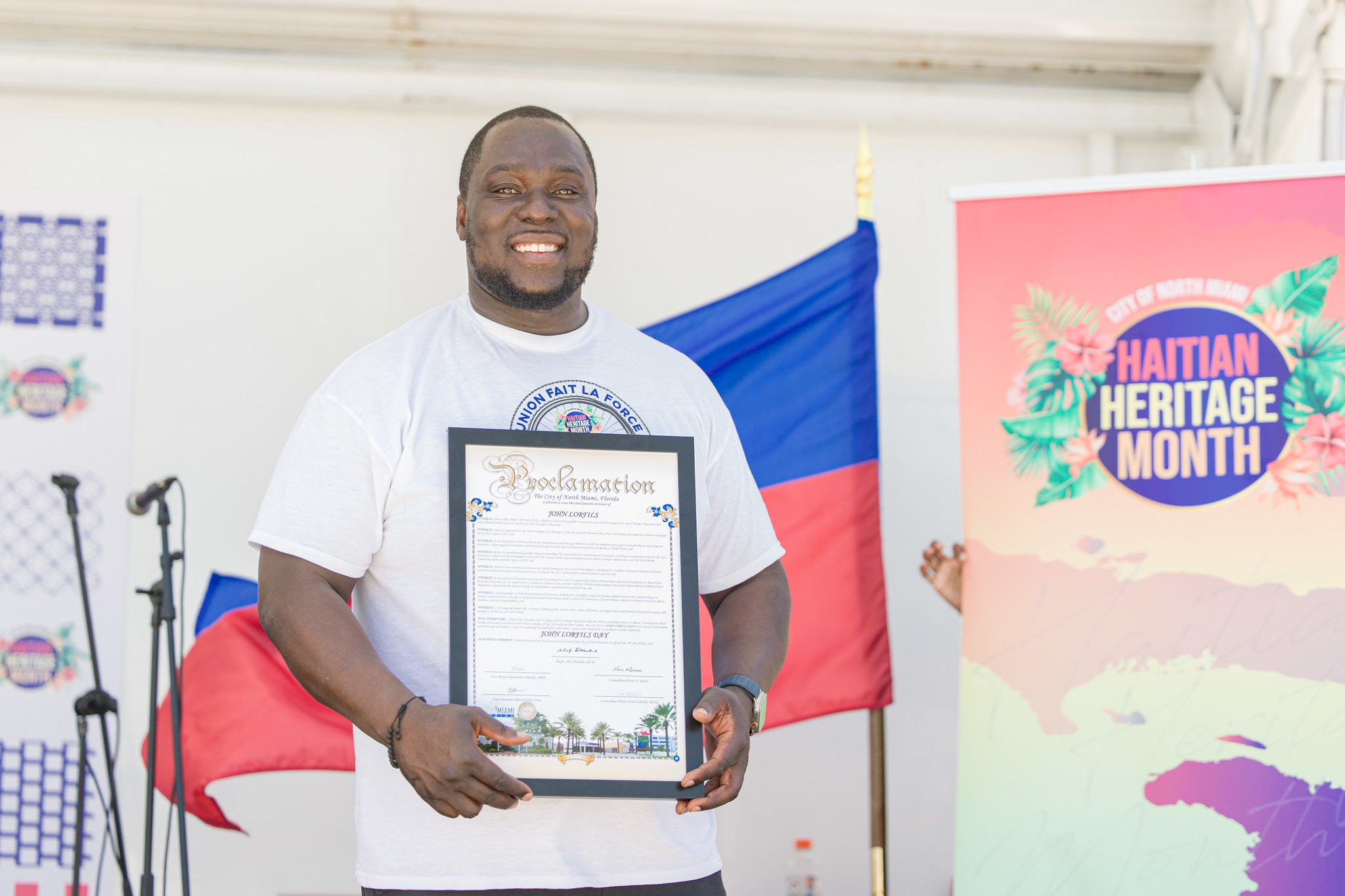 Man holding a framed certificate, smiling, with a Haitian flag and a banner reading 'Haitian Heritage Month' in the background.
