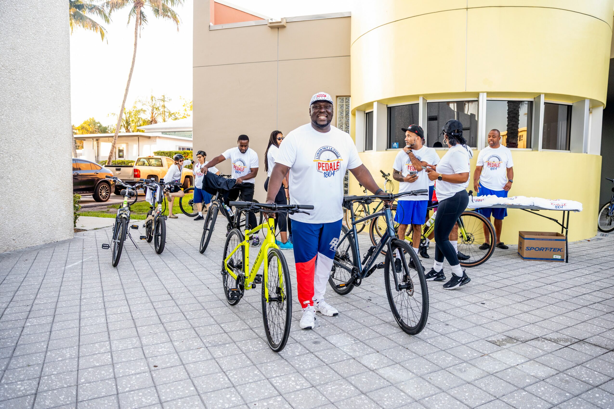 Group of people with bicycles outside a building, some wearing white shirts, smiling and preparing for a ride.