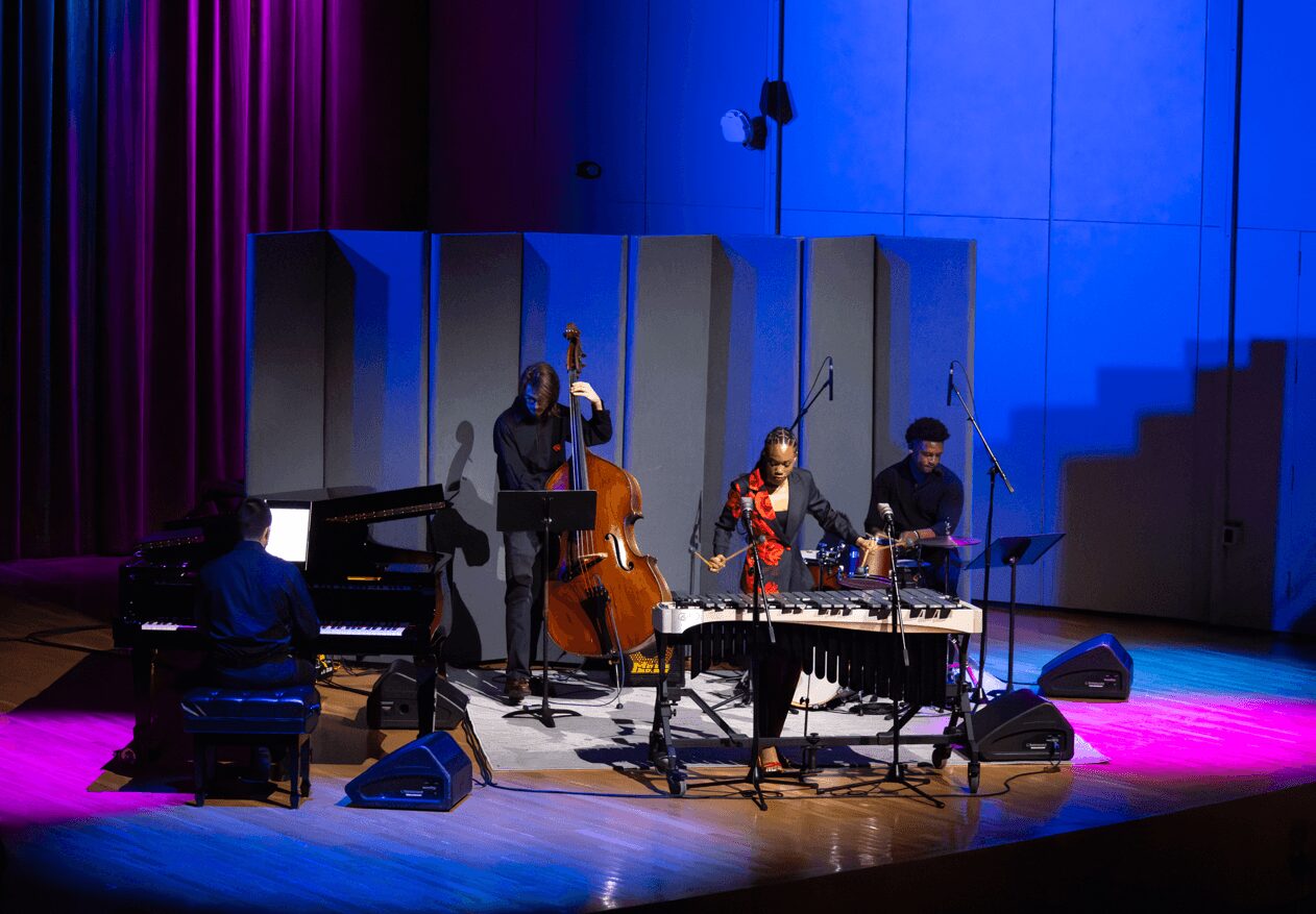 Four musicians perform on stage with instruments, illuminated by blue and purple lighting, in a concert hall setting.