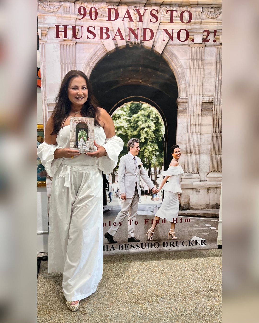 Woman in white dress holding a framed photo, standing in front of an archway with people walking behind her.