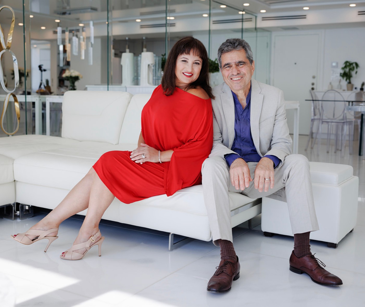 A woman in a red dress and a man in a light suit sit on a white sofa in a modern, bright room.