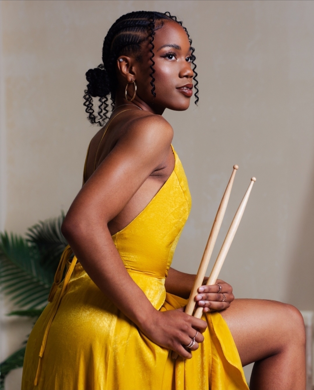 Woman in yellow dress holding drumsticks, sitting with one leg raised, against a plain background.