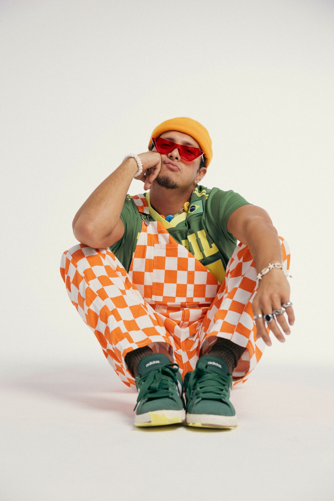 Person sitting on the floor wearing colorful checkered pants, green shoes, yellow beanie, and red sunglasses, with a neutral background.