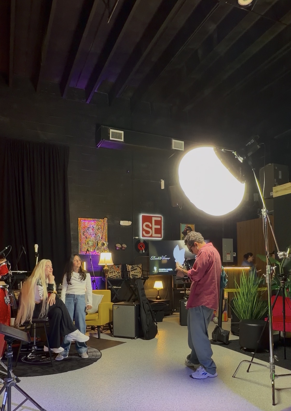 People sitting and standing in a room with colorful furniture and artwork, a large bright light, and a person in a red jacket.