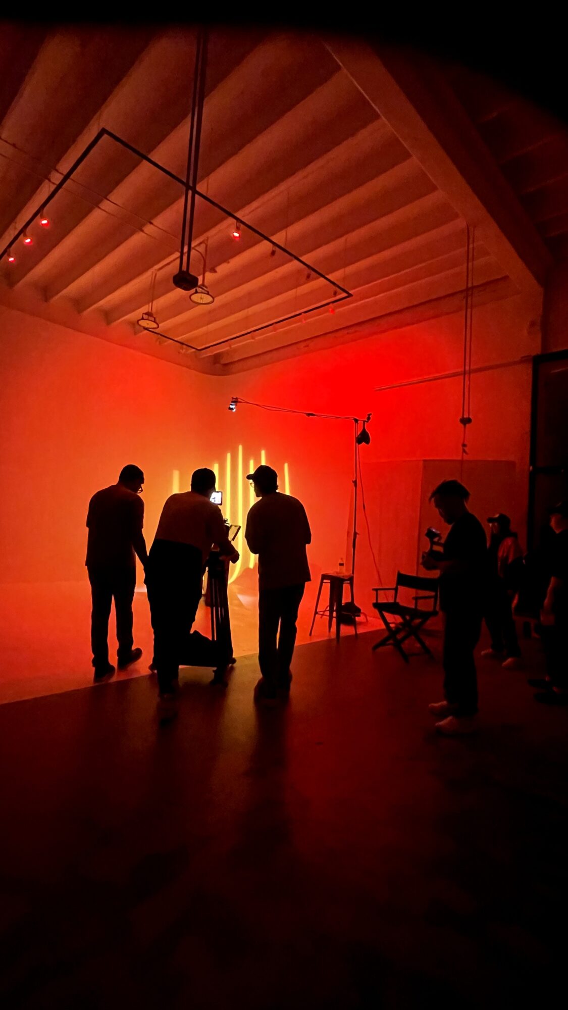 Silhouettes of people in a dimly lit room with red lighting, some standing and talking, with equipment and a high ceiling.