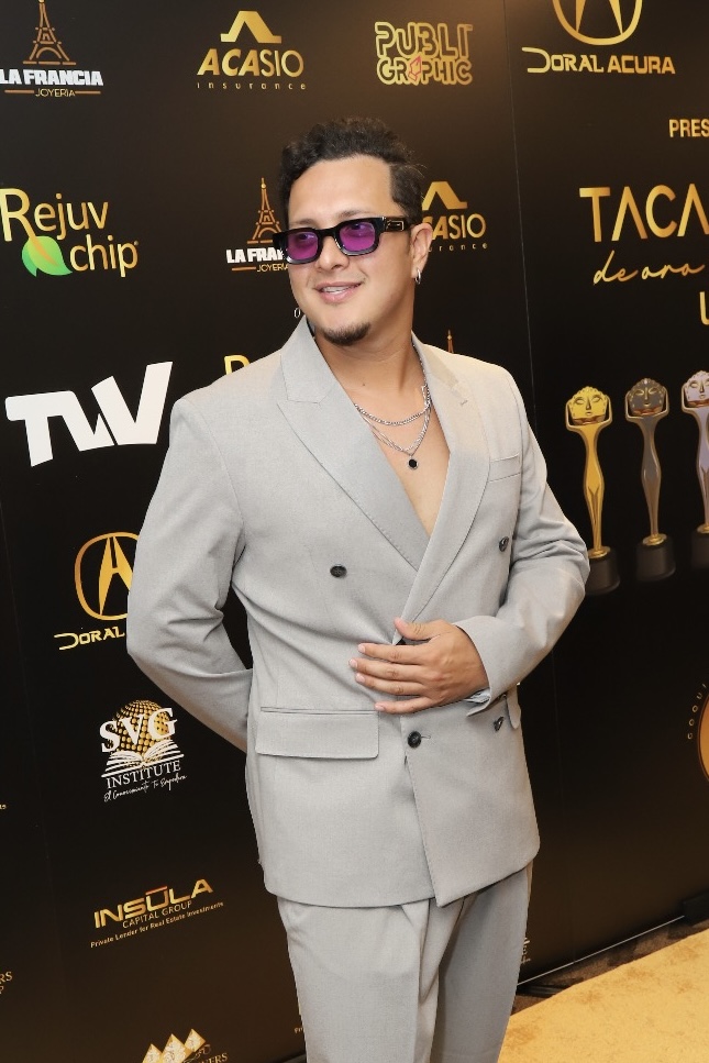 Person in a light gray suit with sunglasses, standing in front of a black backdrop with logos and awards, smiling.