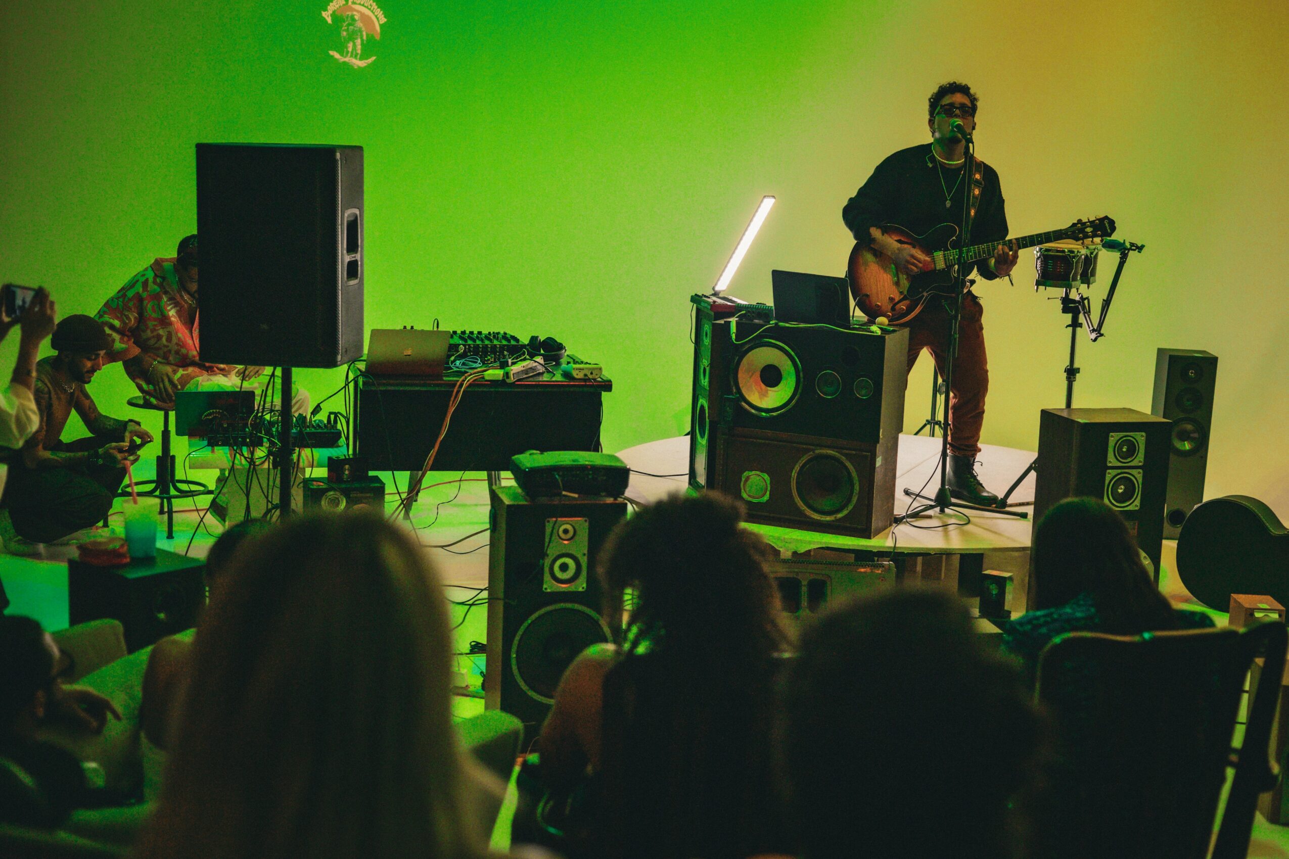 Musician playing guitar on stage with speakers and colorful lighting, audience watching in foreground.