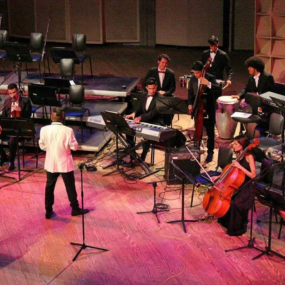 Orchestra performing on stage with conductor, musicians playing instruments including keyboard, drums, and cello, in a concert hall.