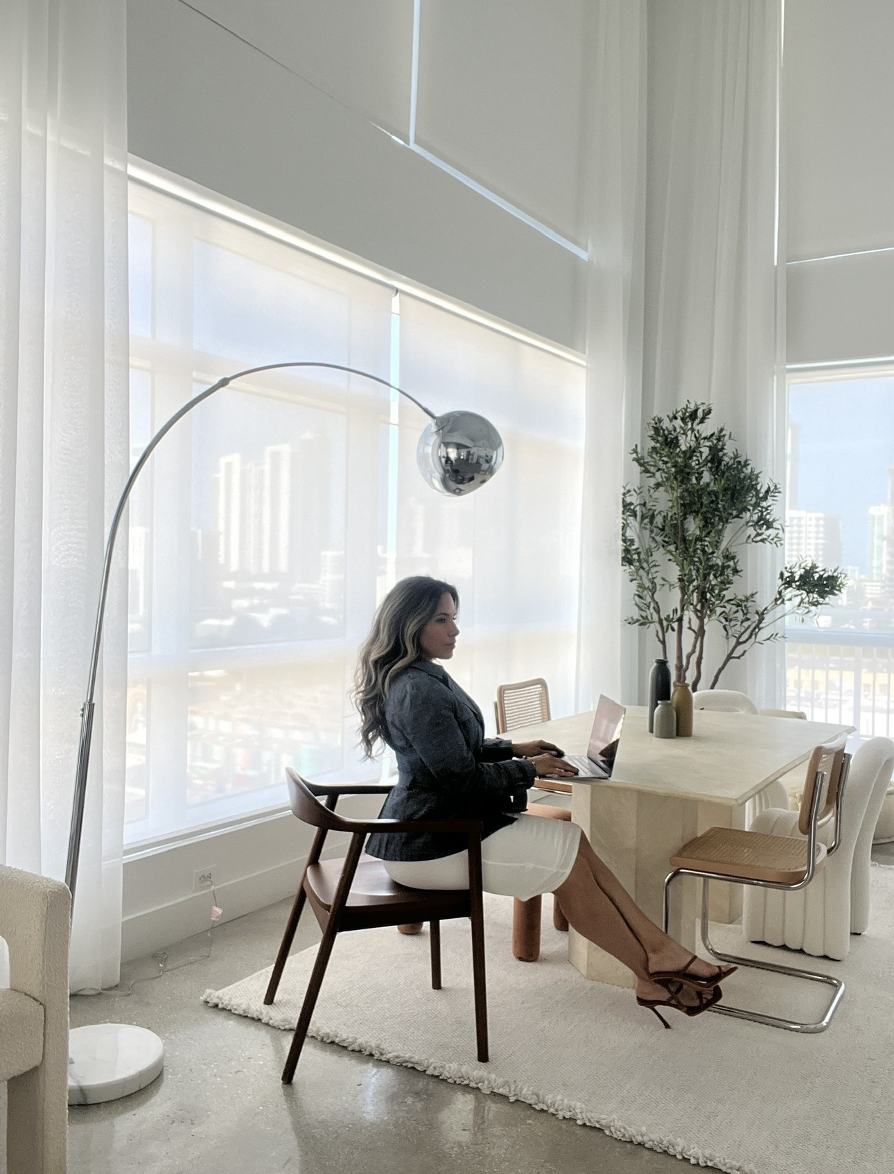 Woman sitting at a dining table with a laptop, in a bright room with large windows and potted plants.