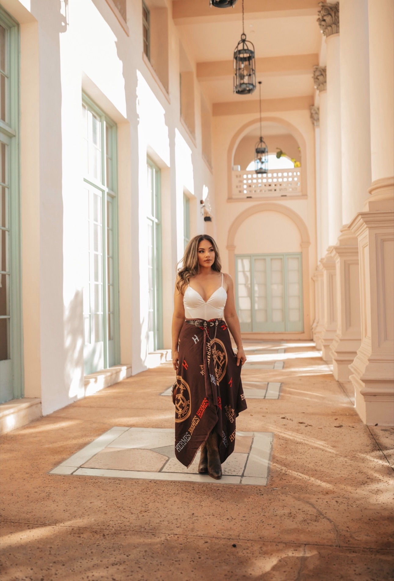 Woman in a white top and patterned skirt walking in a sunlit corridor with tall windows and arches.