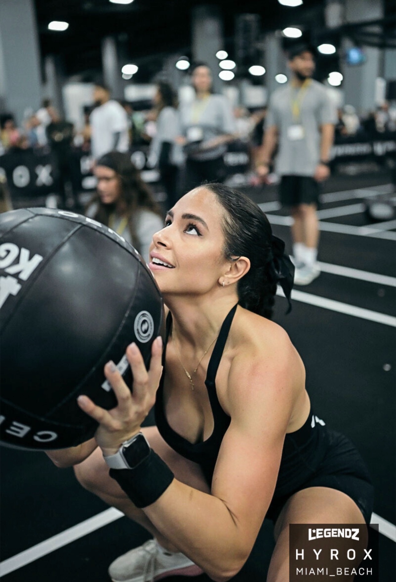 Woman holding a large black exercise ball in a gym, smiling, with people working out in the background.