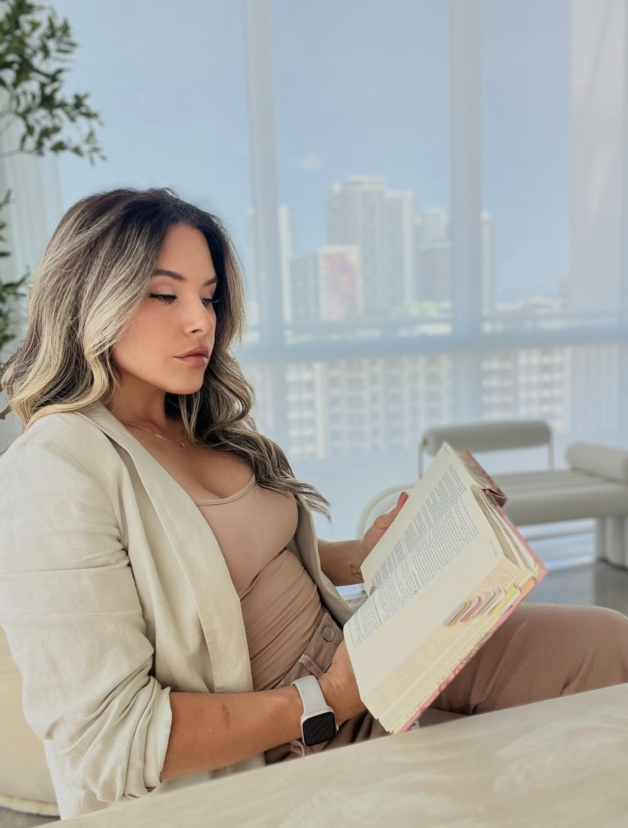 Woman sitting indoors reading a book, with city buildings visible through large windows behind her.