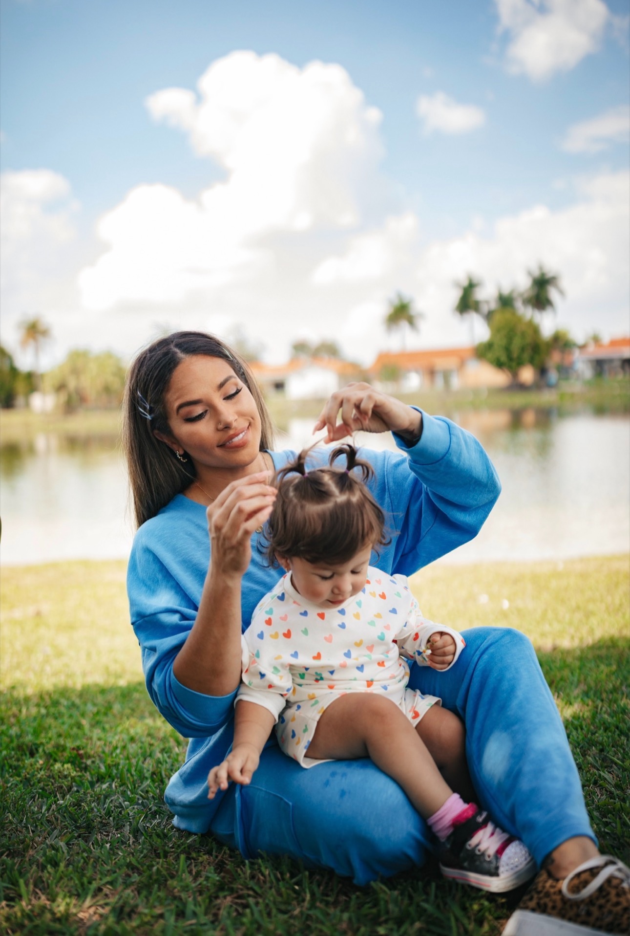 Woman sitting on grass by water, holding a child's hair, outdoors with trees and buildings in background.