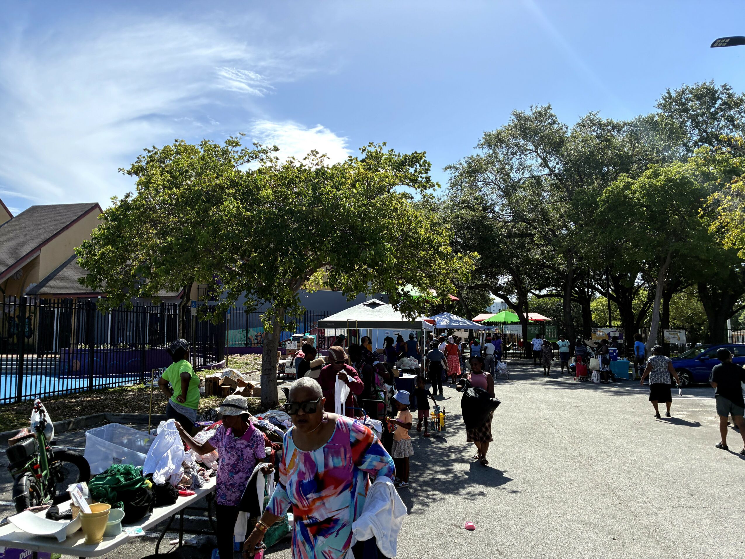 People gather outdoors under trees, some sitting at tables, others walking, with a blue sky and buildings in background.