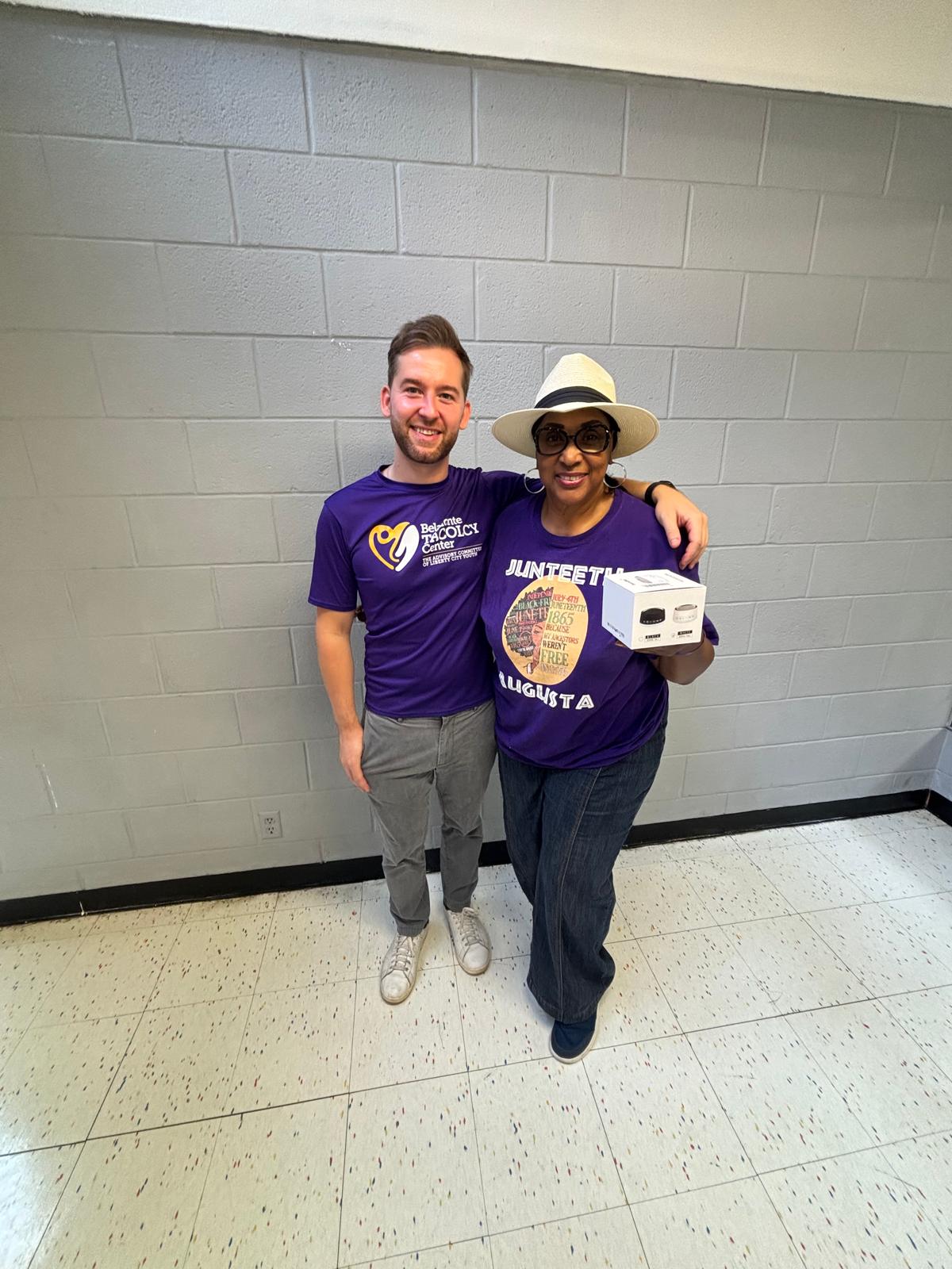 Two people standing against a gray wall, smiling, one holding a box, both wearing purple shirts, one with a hat.