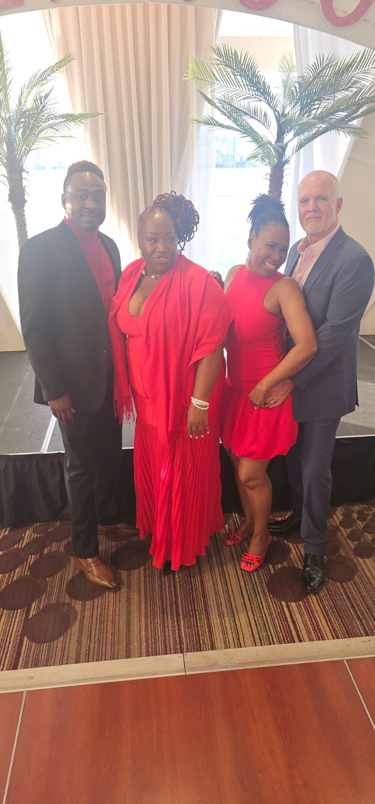 Four people standing together indoors, two women in red dresses, two men in dark suits, with palm trees and curtains behind.