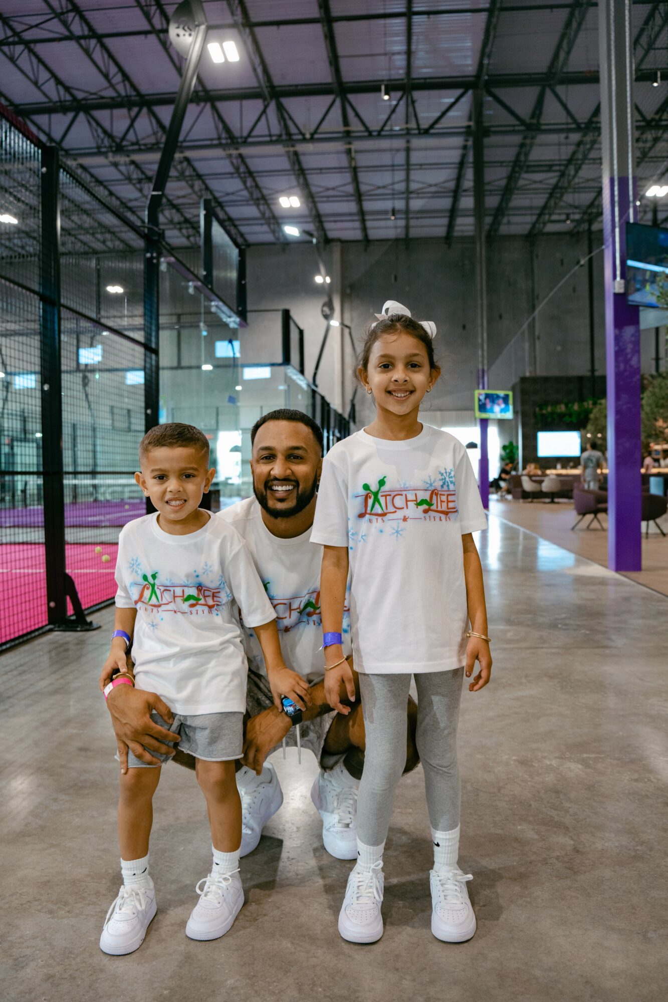 Man with two children in an indoor sports facility, all wearing white T-shirts and sneakers, smiling at the camera.