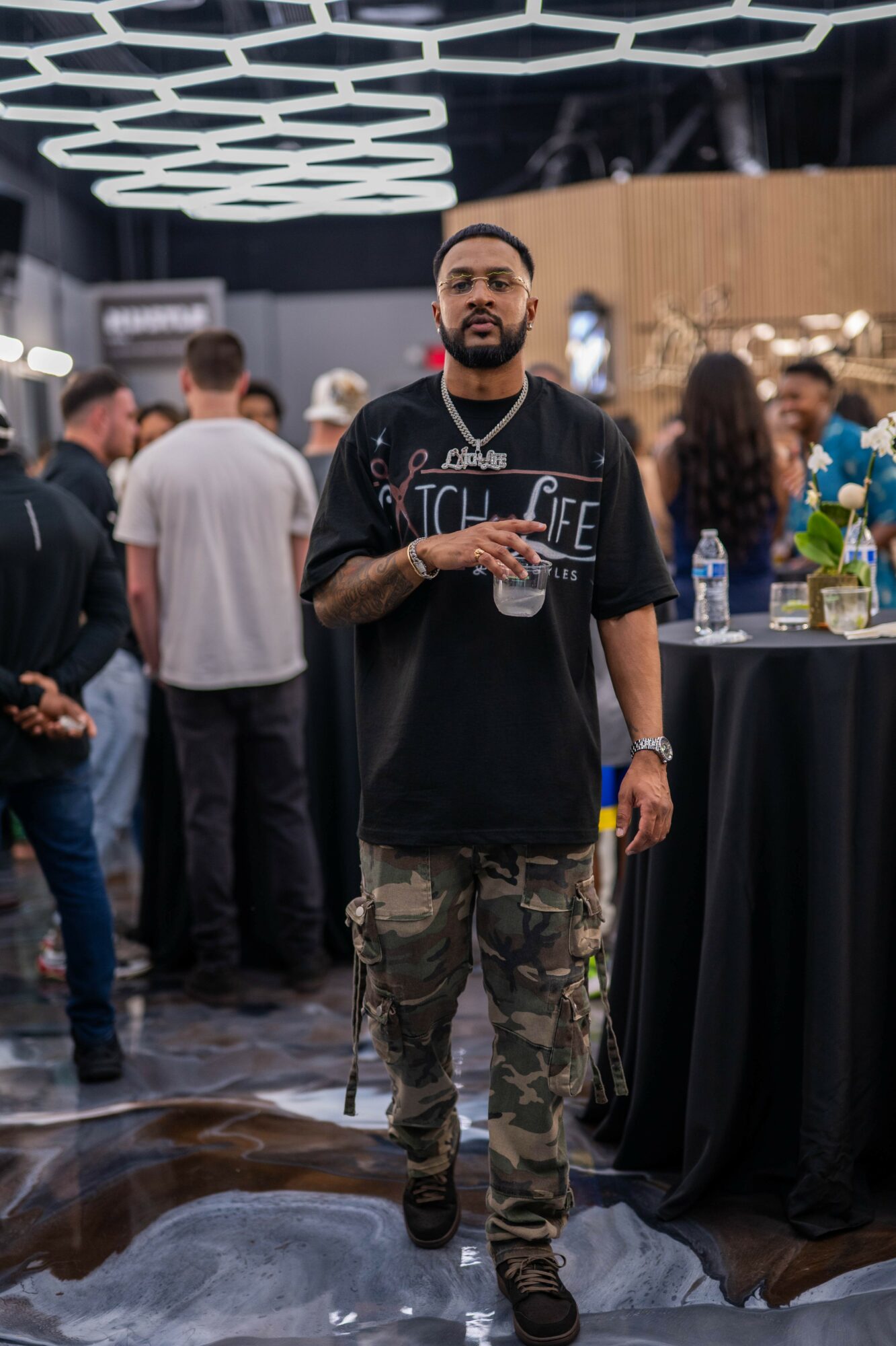 Man standing at a social event holding a drink, with people and a bar in the background.