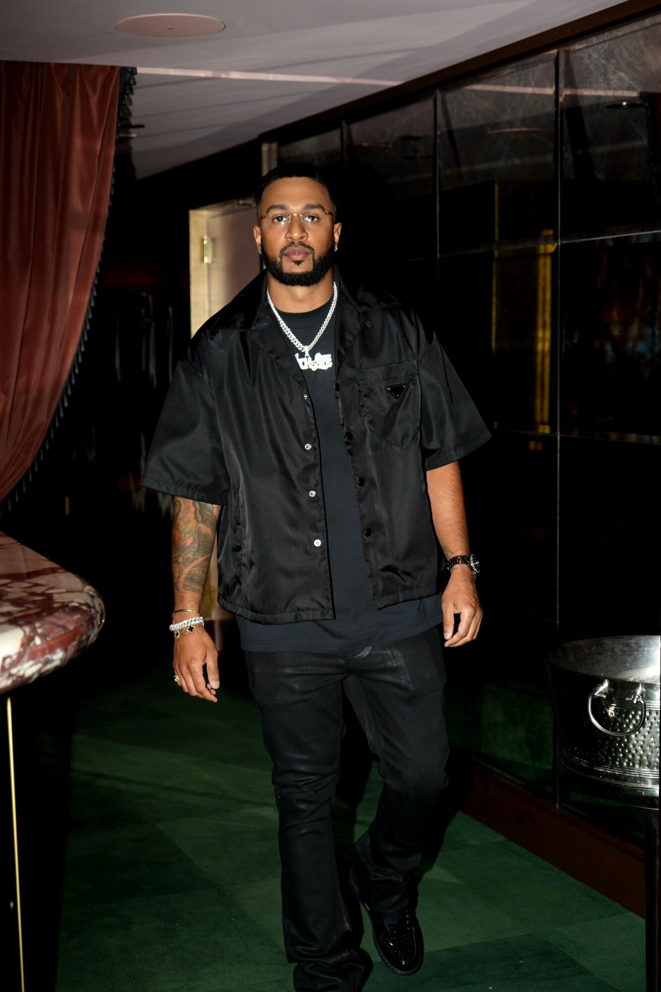 Man with tattoos and jewelry stands indoors, wearing black clothing and a chain, with dark background and curtains.