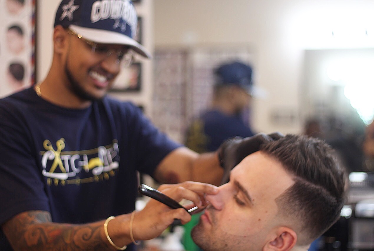 Barber using a marker on a client's face in a barbershop, smiling, with other clients in the background.