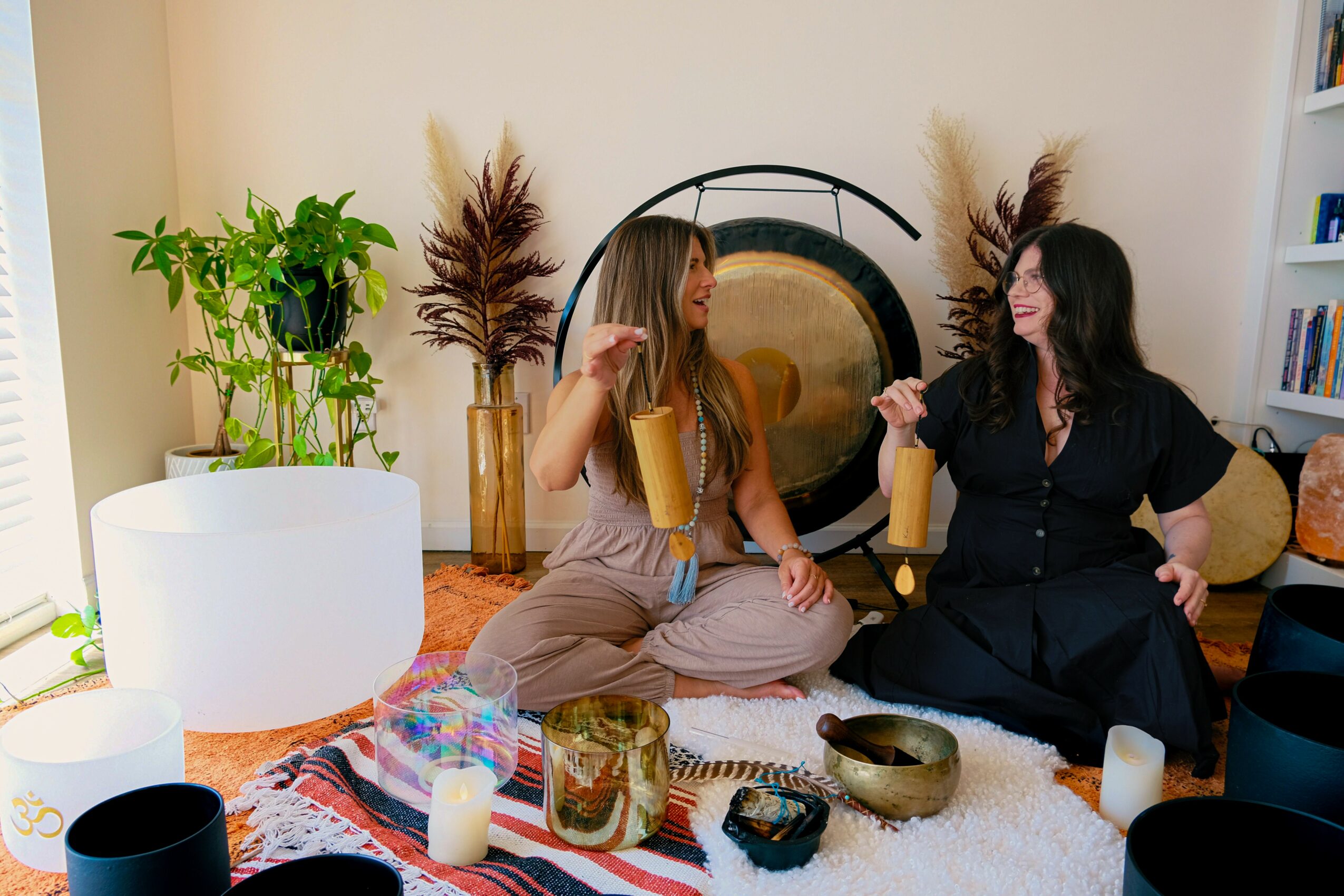 Two women sit on a rug in a cozy room, talking and smiling, surrounded by plants, candles, and decorative objects.