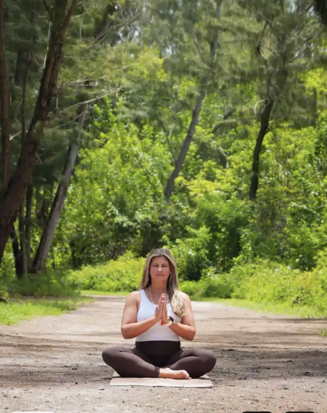 Person sitting cross-legged on a mat in a forest, practicing meditation with hands in prayer position, surrounded by green trees.