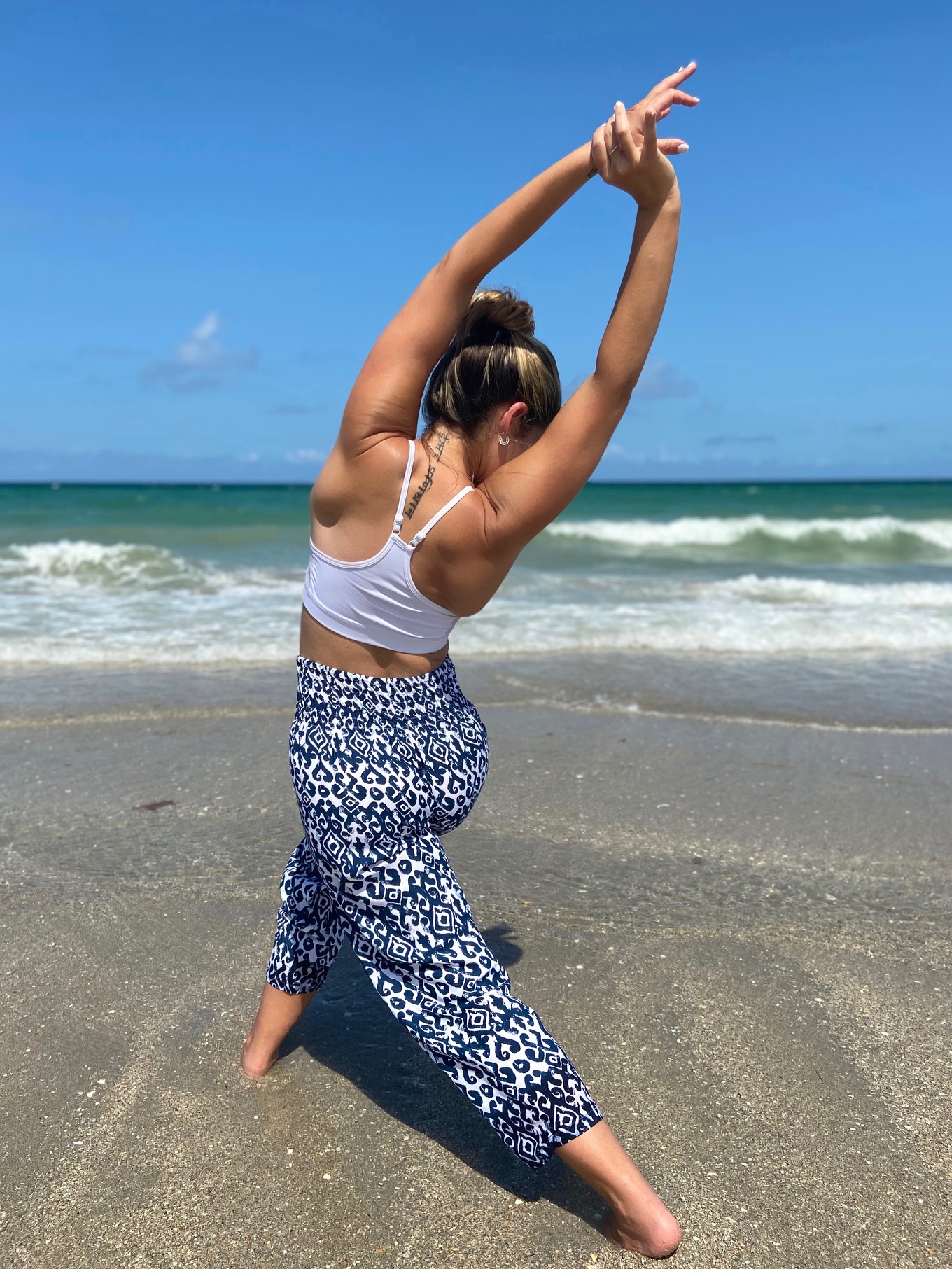 Person practicing yoga on the beach with arms raised, facing away from the camera, ocean and sky in background.