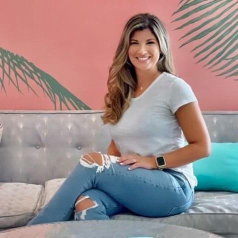 Woman sitting on a sofa, smiling, with a pink wall and green plant shadows in the background.
