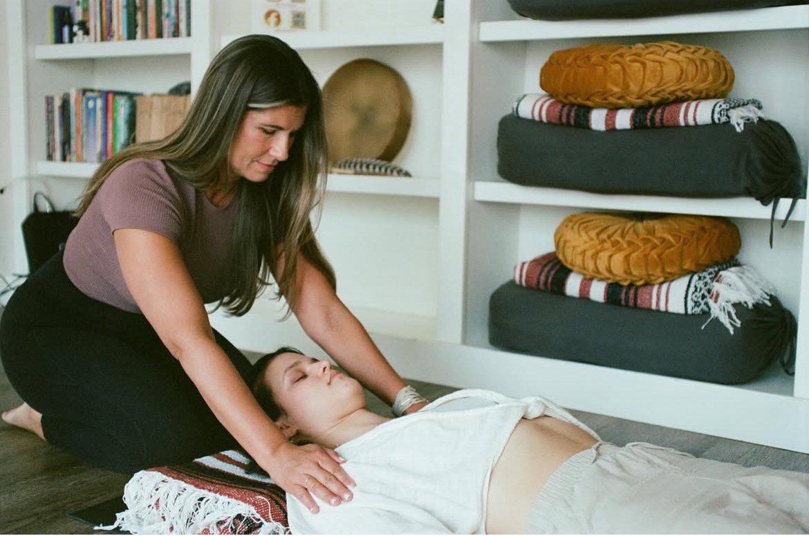 Woman performs a massage on a person lying on the floor in a room with shelves and folded blankets.