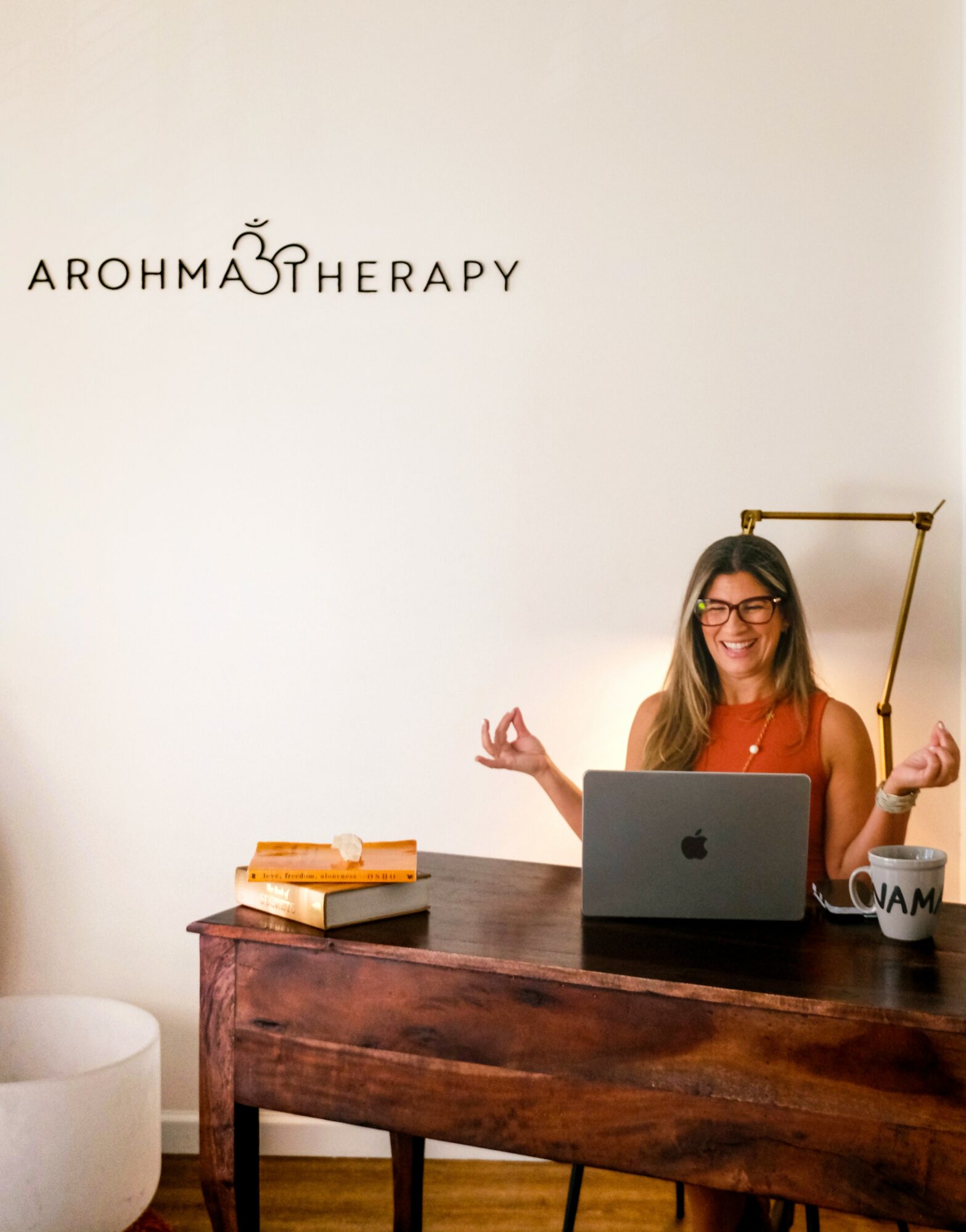 Woman sitting at desk with laptop, smiling, holding a lamp, with books and a mug on desk, and a sign on wall.