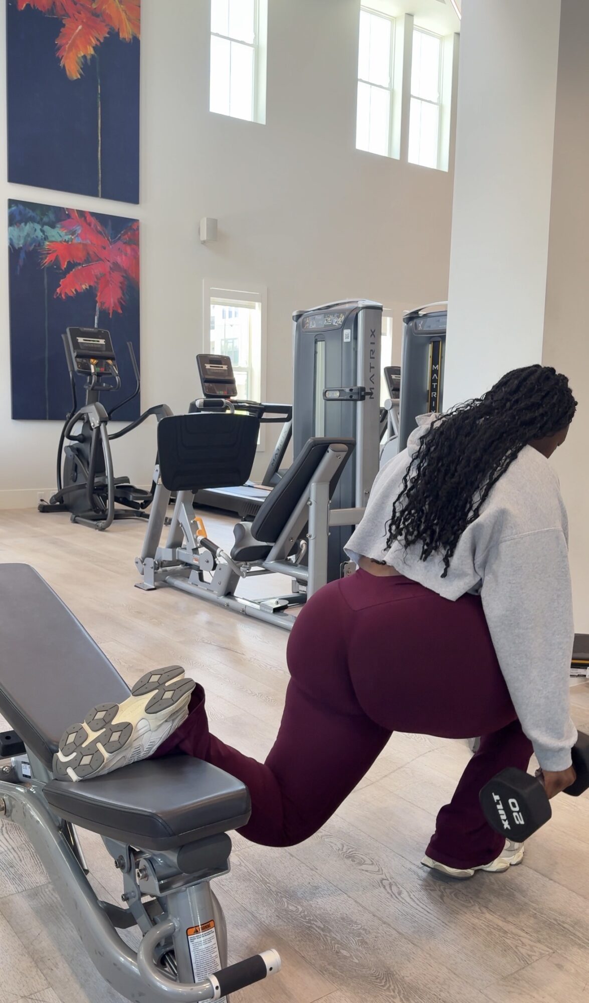 Woman in gym using dumbbell, exercise equipment in background, large windows, modern interior, light-colored walls.