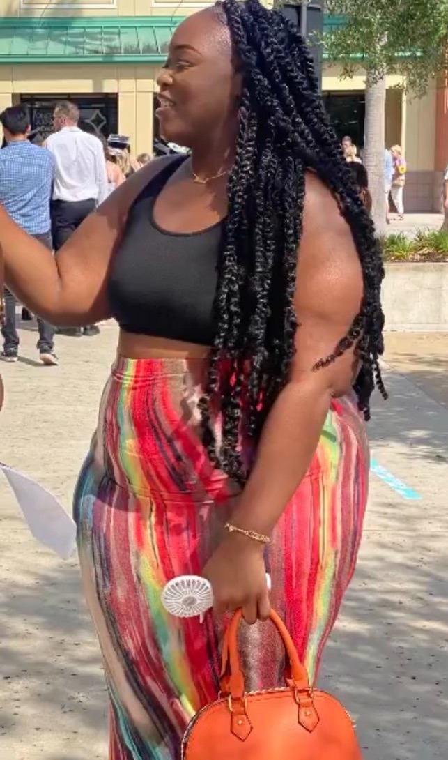 Smiling woman with long braided hair wearing a black top and colorful striped pants, holding a small orange bag and a fan.