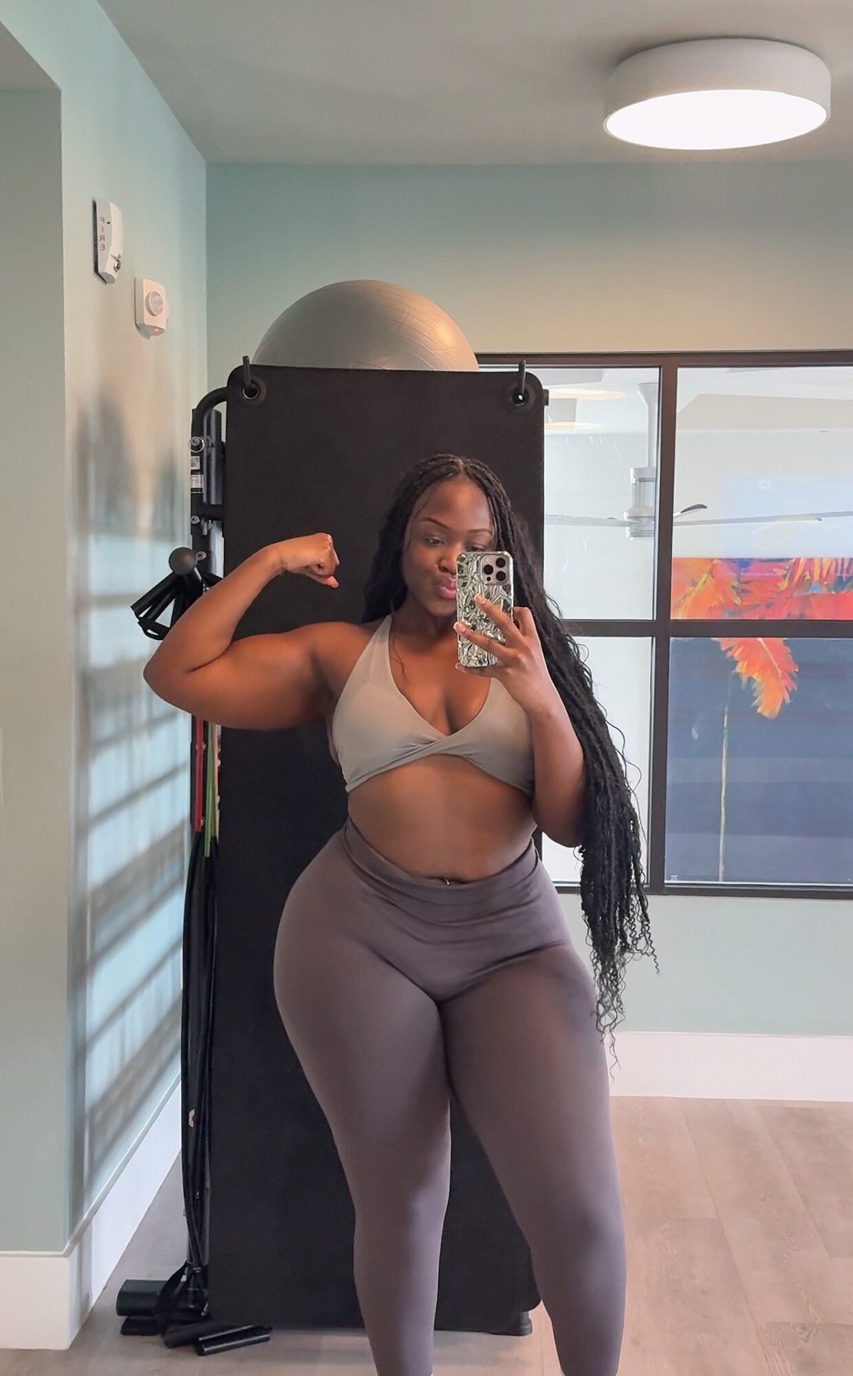 Woman taking a mirror selfie in a room with large window, wearing sports bra and leggings, flexing arm muscle.