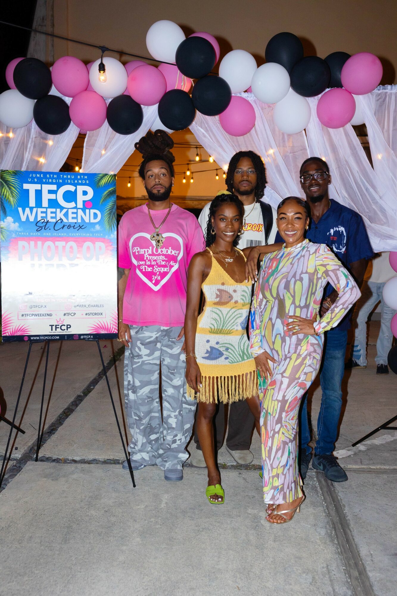 Group of five people standing under decorated arch with pink, black, and white balloons at event entrance.