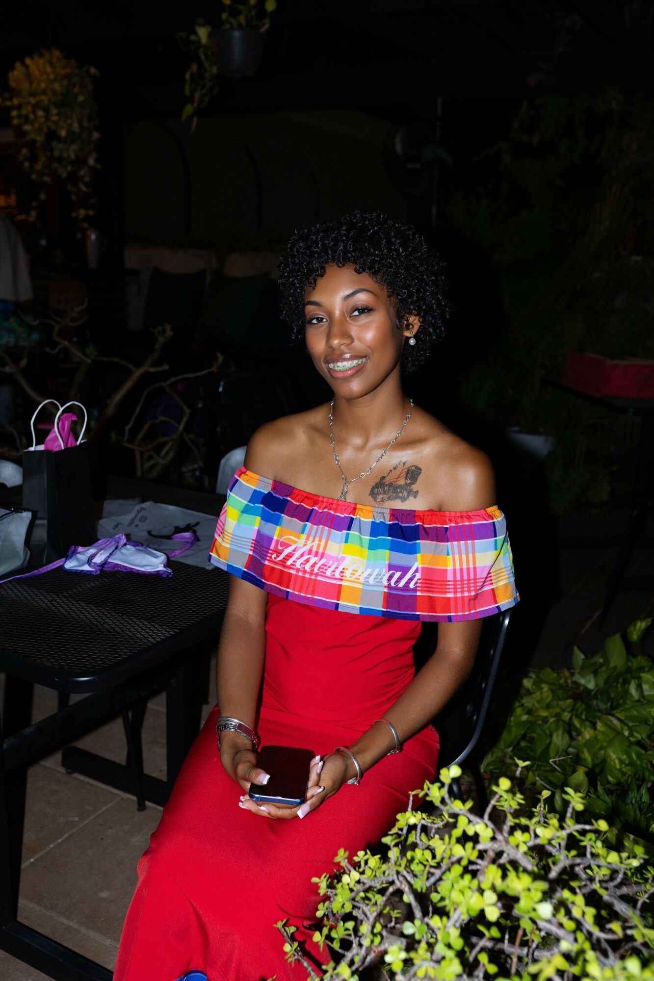 Woman in red dress with colorful off-shoulder top, holding a phone and a clutch, smiling outdoors at night.