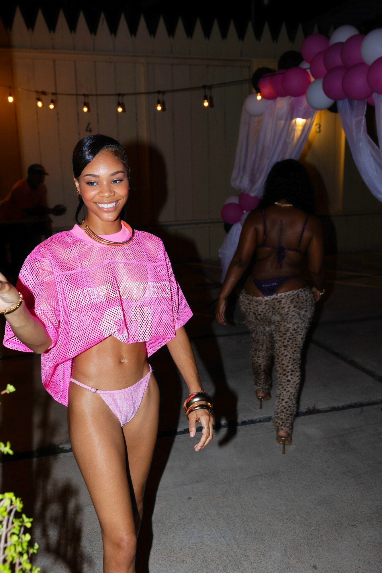 Young woman in pink crop top and underwear smiling at party, decorated with pink balloons and string lights.