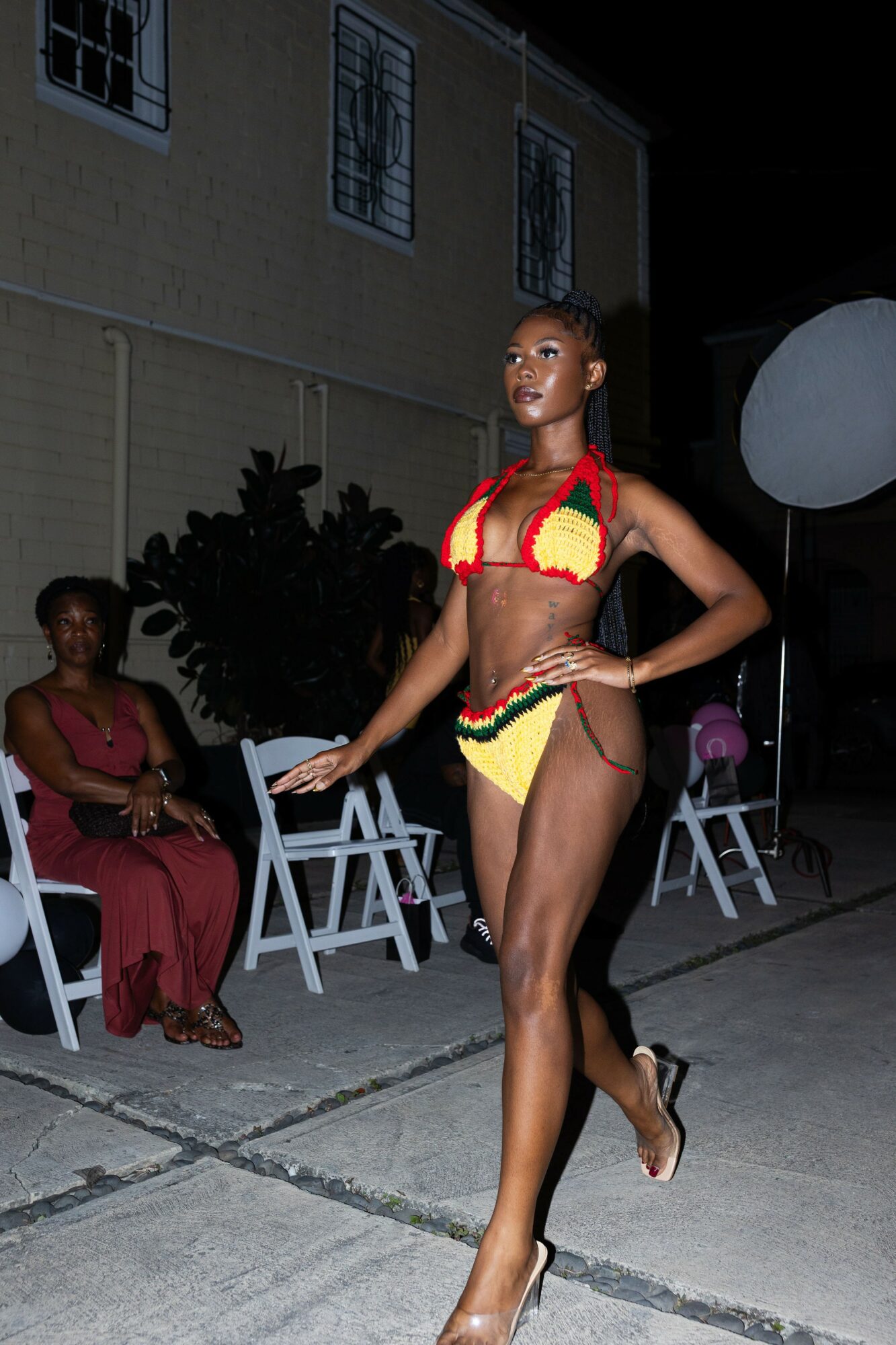Young woman in colorful bikini walking outdoors at night, with seated woman in red dress nearby, and chairs and plants in background.