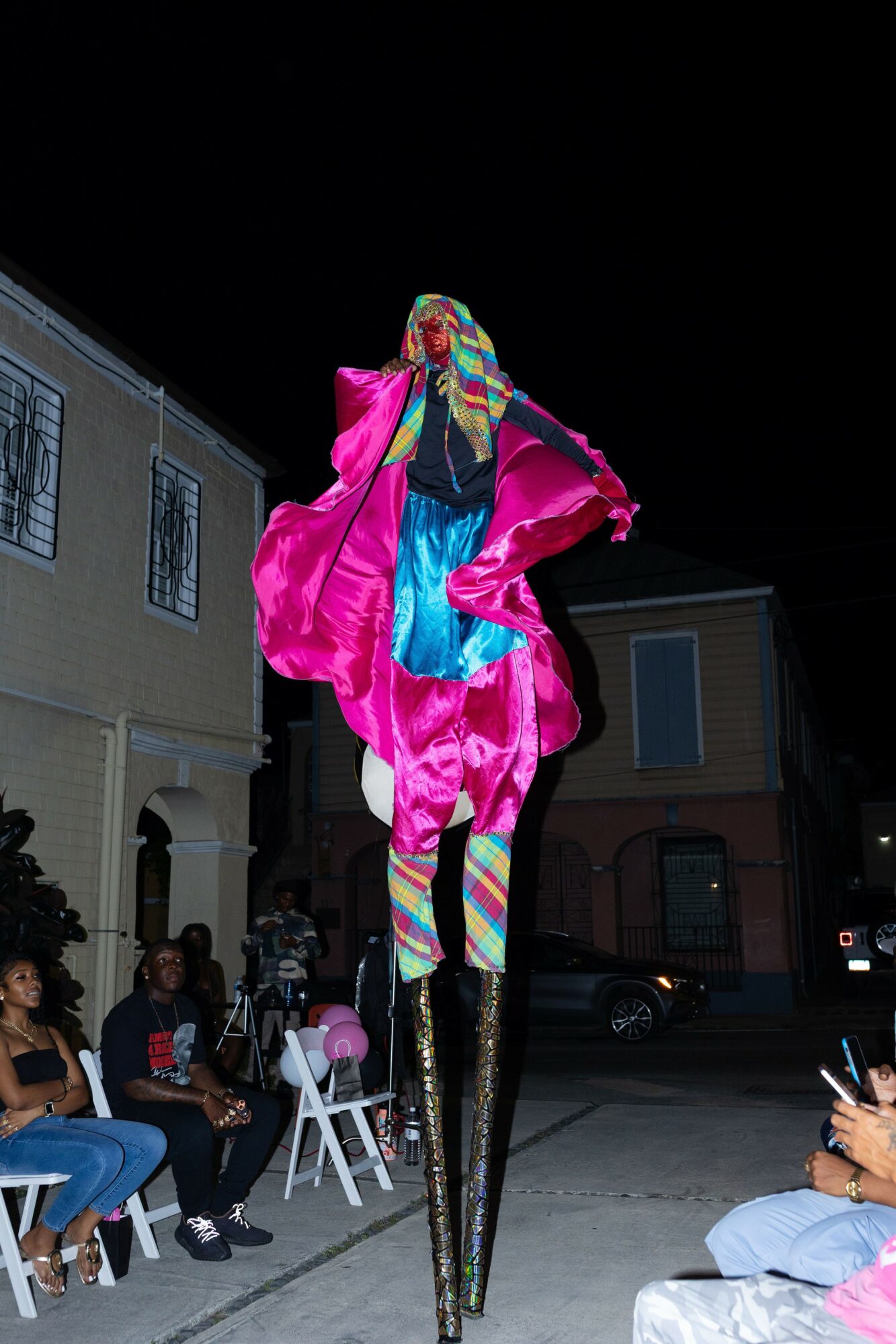 Person dressed in colorful costume with tall stilts, pink coat, and rainbow-striped leggings, standing outdoors at night.