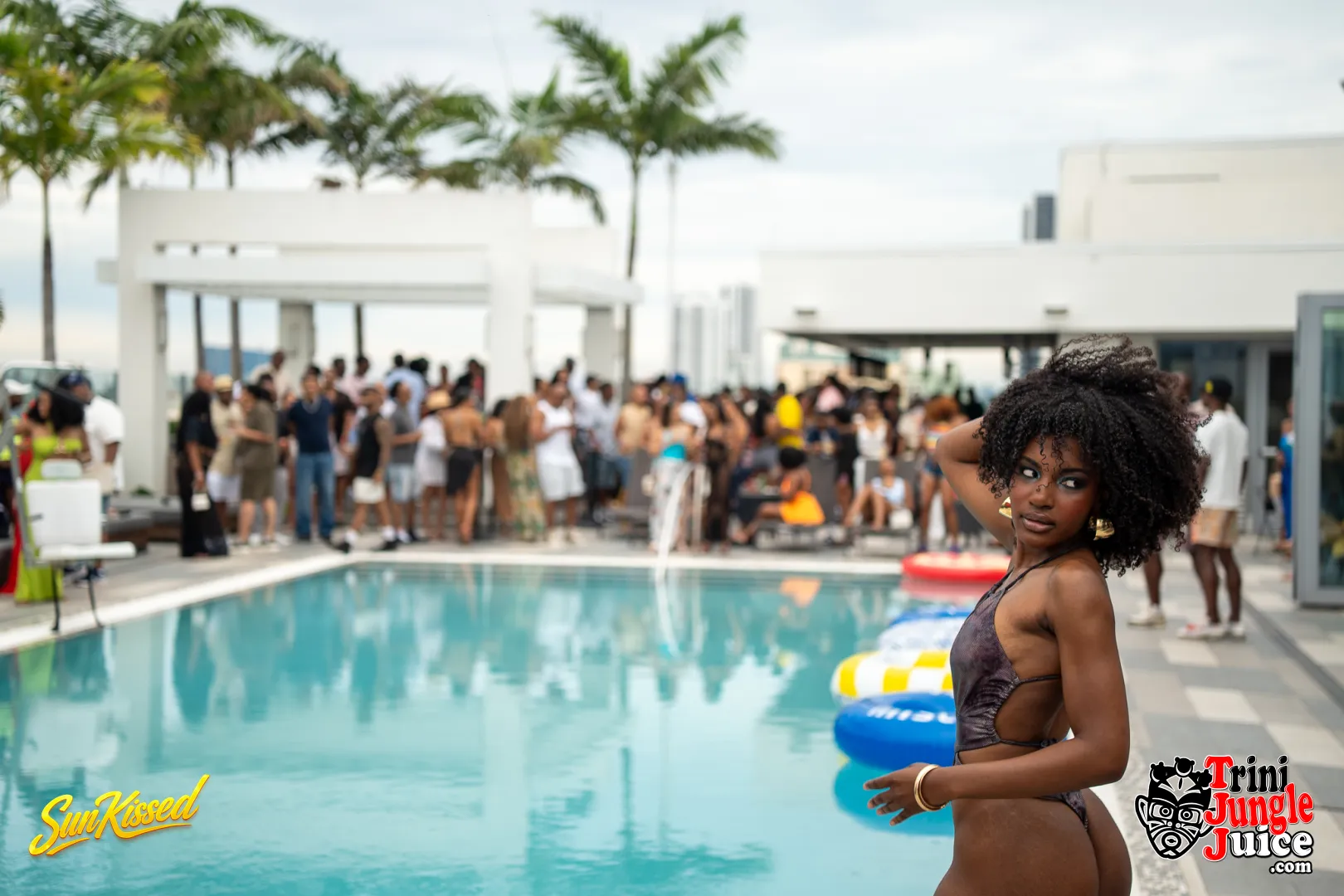 Young woman with curly hair standing by a pool with people in the background, palm trees, and a modern building.